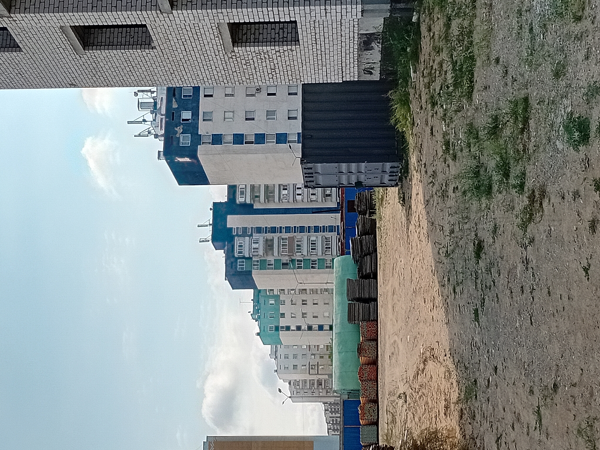 In the city of Semey, Kazakhstan, a construction site is depicted with multiple apartment buildings in various stages of completion. On the right, a partially constructed building made of light-colored bricks displays open window frames. Adjacent to it is a dark grey shipping container. In the background, a row of completed multi-story apartment buildings stretches from right to left, characterized by their blue, white, and green facades. Some of these buildings have visible antennas on their roofs. In the foreground, stacks of building materials are visible, including what appear to be rolled-up green tarps or insulation, and stacks of dark grey slabs, possibly concrete or stone. The ground is a mixture of dirt and sparse vegetation. The sky is a light blue with scattered white clouds.