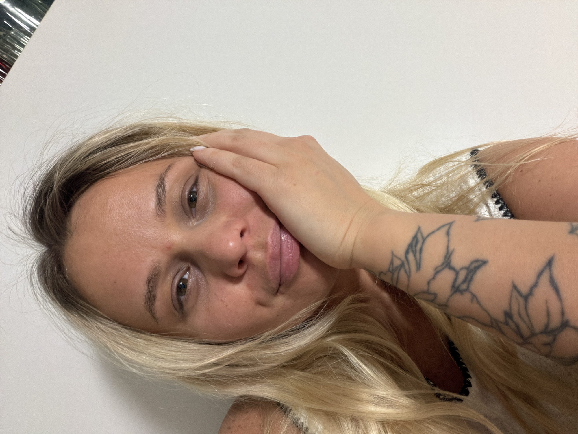 A woman with light blonde hair is holding her hand to her face. The woman has tattoos on her arm. The background appears to be a plain white wall. There is no discernible information that indicates that this image was taken in Sarapul, Russia.