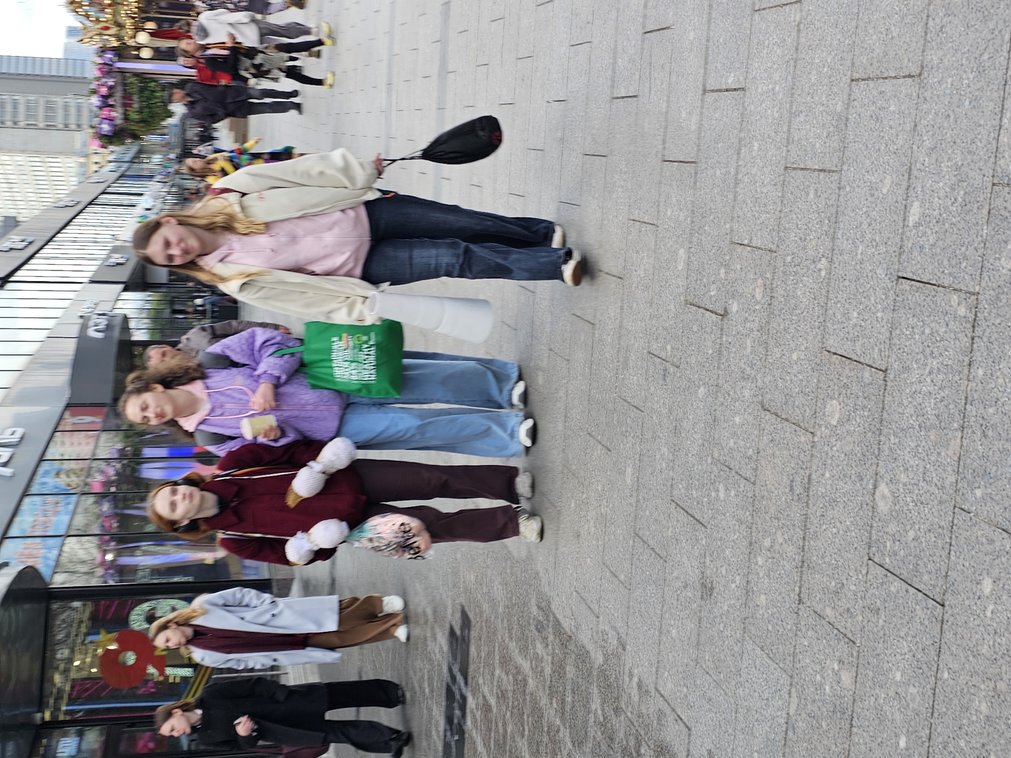 The image shows a group of people on a sidewalk in Moscow, Russia. There are four girls lined up, while others walk past. The first girl is wearing a pink sweatshirt and a cream jacket with blue jeans. The next girl is wearing a purple sweatshirt and jeans, and is carrying a green shopping bag. The third girl is wearing a maroon jacket and pants. The fourth girl has on a brown pair of pants with a grey jacket and red scarf. Behind the fourth girl is a person in a black suit. The sidewalk is paved with grey, rectangular stones. In the background is a glass building with storefronts, and buildings in the distance. There are a lot of people on the sidewalk.