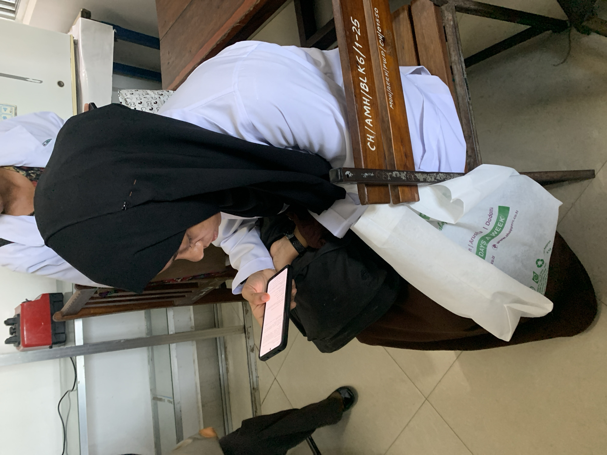 The image shows a person wearing a black head covering and a white top, sitting on a wooden chair while looking at their mobile phone. The chair has inscriptions "CH/AMH/BLK6/1-25" and "MOH/ARRH/PWD/CH/01550" on its wooden frame. Beside the person is a white paper bag with the words "Arusha | Dodoma | DAYS A WEEK | www.shoppers.co.tz". The floor is tiled. The overall setting appears to be indoors, possibly a waiting area. This is in Tanzania, a city with the city of Arusha as one of its regions.