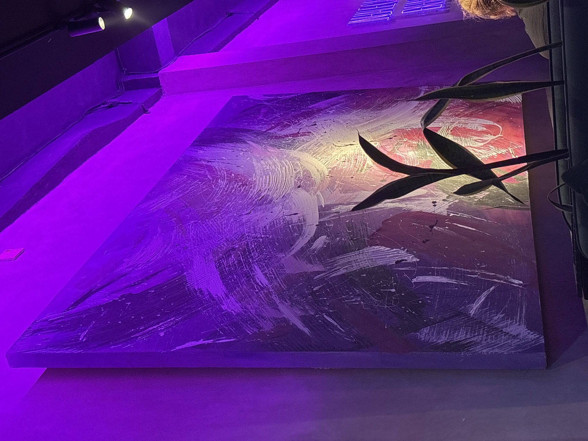 A large, abstract painting with bold, swirling brushstrokes is mounted on a wall under purple lighting. The painting is a mix of white, red, and purple hues. To the right of the painting, there are several tall, dark green plants with sharp, pointy leaves. The scene appears to be indoors. There is no information about the location in Kuta Selatan, Indonesia.