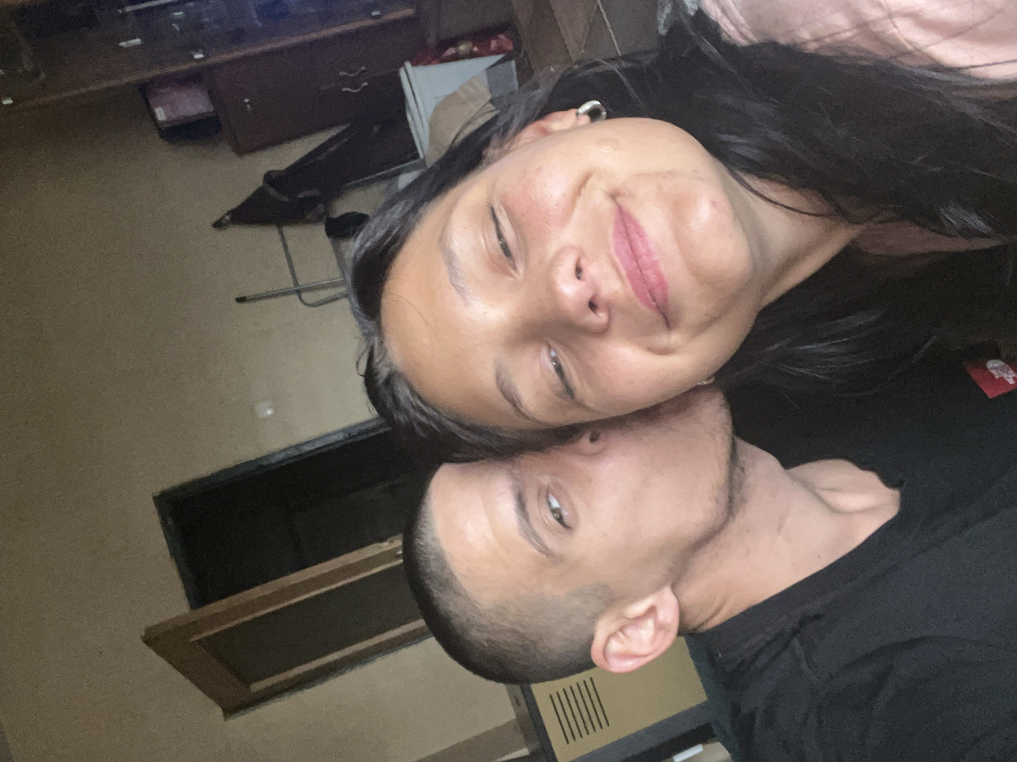 A selfie captures a couple in an indoor setting. The woman, with dark hair and a pink top, smiles at the camera, while the man, wearing a black shirt, is seen from the side, his head close to hers. The background reveals a room with furniture and a cabinet. There is no information in the image to determine it was taken in Sofia, Bulgaria.