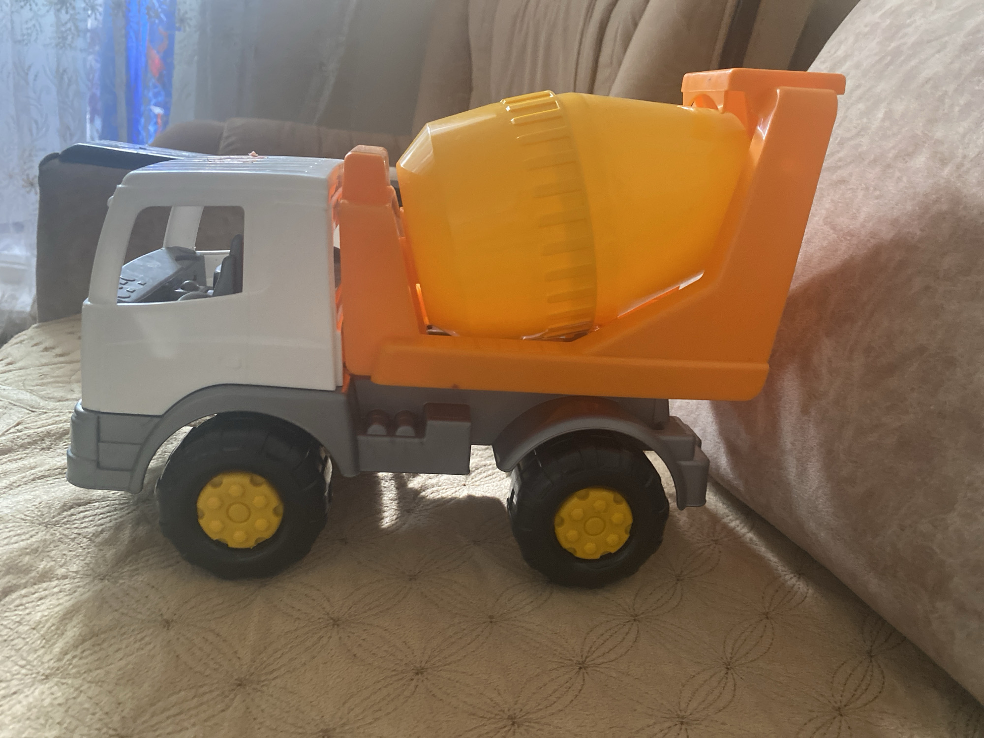 The image shows a toy cement mixer truck, predominantly orange and yellow with a white cab, resting on a textured beige surface that appears to be upholstery. The truck is positioned on its wheels, with the drum of the mixer facing towards the right of the frame. The background is softly blurred, suggesting a domestic environment. A hint of a couch and curtains can be discerned. There is no information in the image to indicate a location in Kizilyurt, Russia.
