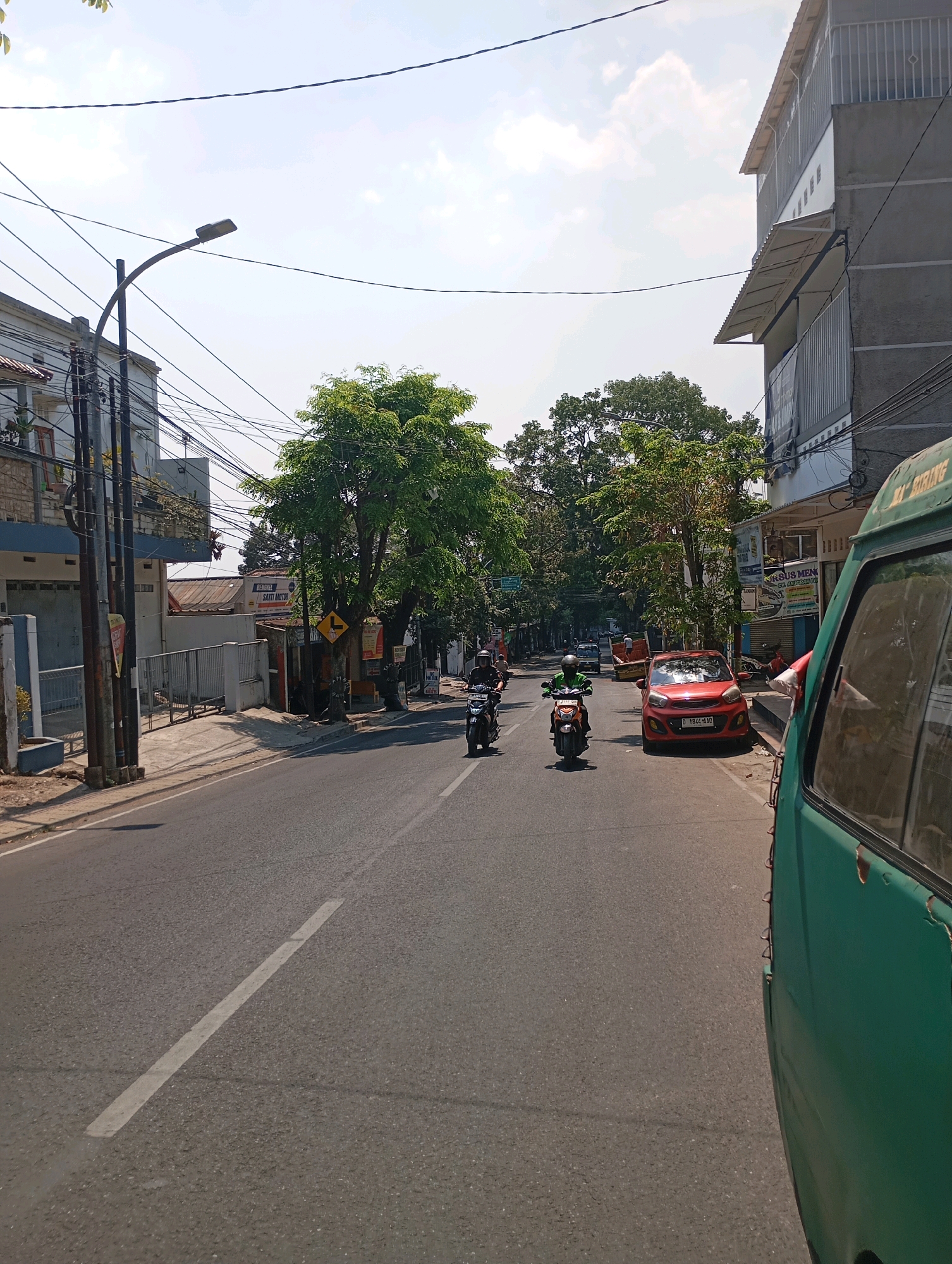 The image depicts a street scene in Cimahi, Indonesia, on a clear, sunny day. A paved road occupies the majority of the frame, with a white dashed line marking the center. Several motorcycles and a red car are visible on the road. On the left side of the road, there is a sidewalk with a white fence and buildings with multiple stories. A street lamp is also present on the left. The right side of the road features more buildings, some with awnings, and a green vehicle partially obscures the view. Lush green trees line the street, suggesting a somewhat urbanized but also verdant environment. The sky is bright blue with some scattered white clouds.