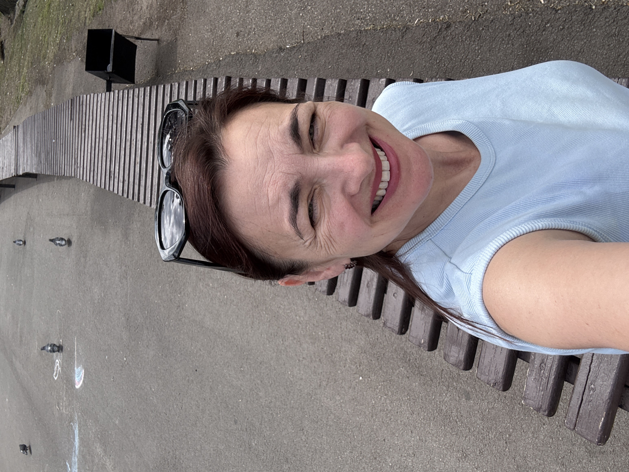 A woman is sitting on a long, wooden bench outdoors. She is wearing a light blue top and sunglasses are perched on top of her head. She is smiling widely, with her eyes closed. The bench is set on an asphalt surface, and a trash can is visible in the background. Several pigeons are scattered on the ground in front of the bench, and some chalk drawings are also visible on the asphalt. The lighting suggests it is daytime, and the weather appears to be overcast or possibly late afternoon. The scene takes place in a park or public outdoor area. There is no visible text in the image.