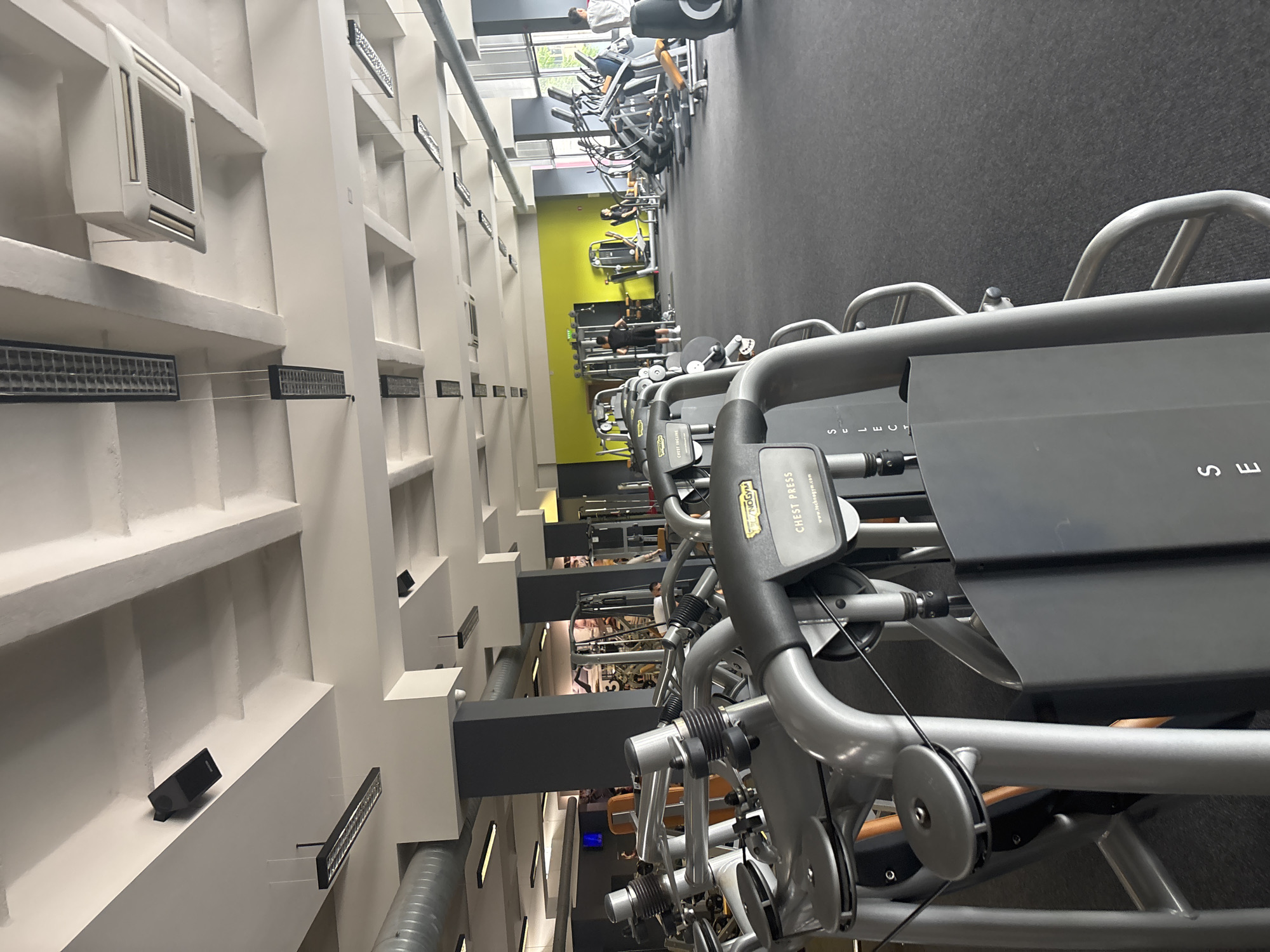 This image captures an indoor fitness center or gym, viewed from an elevated perspective looking down onto the workout floor. The setting appears modern and well-lit, with natural light entering from large windows in the background. The floor is covered in dark grey, likely rubberized material.
In the immediate foreground, several "Technogym" brand "Chest Press" machines are prominently featured, characterized by their grey metal frames, black padded sections, and control panels. Visible text on these machines includes "CHEST PRESS" and "www.technogym.com". Further into the mid-ground, rows of various exercise equipment, including cardio machines like treadmills and other strength training apparatus, are arranged. Several individuals are present, actively engaged in exercising.
To the left, a tall, white architectural structure, possibly a wall or support column, features recessed sections with multiple rectangular ventilation grilles or light fixtures. A larger white air conditioning unit is also mounted on this structure. A vibrant lime green wall segment is visible in the mid-ground, indicating a distinct zone within the gym. The background shows large windows looking out onto some greenery, suggesting it is daytime. The overall impression is a spacious and active gym environment. No specific location cues are visible within the image to identify Blagoevgrad, Bulgaria, beyond the provided context.