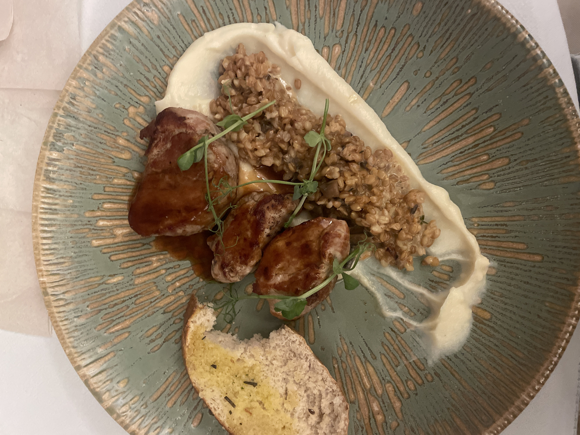 A top-down view of a plate of food, likely in Varna, Bulgaria, displaying an artful arrangement of meat, mashed potatoes, grain, and bread. The meat appears to be pan-fried, glistening with a glaze or sauce. The mashed potatoes form a creamy white smear. The grain mixture is golden-brown and speckled, suggesting a blend of textures and flavors. A partially eaten slice of bread, glazed with a yellow substance and sprinkled with dark specks, sits on the plate's edge. The plate itself is patterned with a radial design in shades of green and brown. A few sprigs of green sprouts garnish the meat and grain. The plate sits on a white tablecloth or placemat. The composition suggests a restaurant setting.