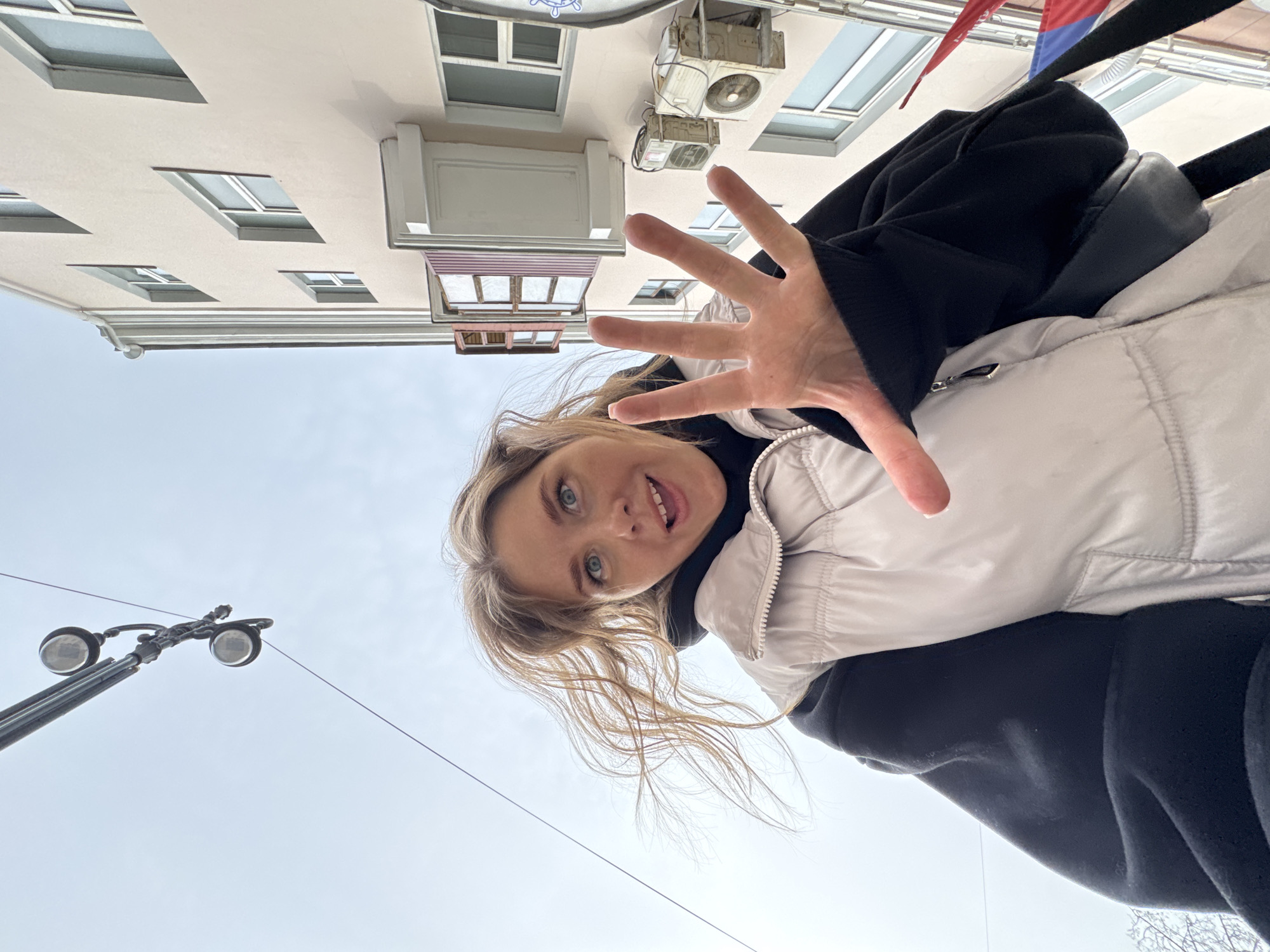 A young woman is visible in the foreground, seemingly taking a selfie from a low angle. She has blonde hair and blue eyes, and is wearing a light-colored padded jacket over a black hooded sweatshirt. Her right hand is extended towards the camera with fingers splayed.
Behind her, a multi-story building with a light-colored facade is visible. The building features multiple windows of varying sizes, some adorned with awnings. Attached to the building are air conditioning units. Above the building, the sky is visible. A street light with two globe fixtures is visible in the lower left corner of the frame. The image was taken in the city of Perm, Russia.