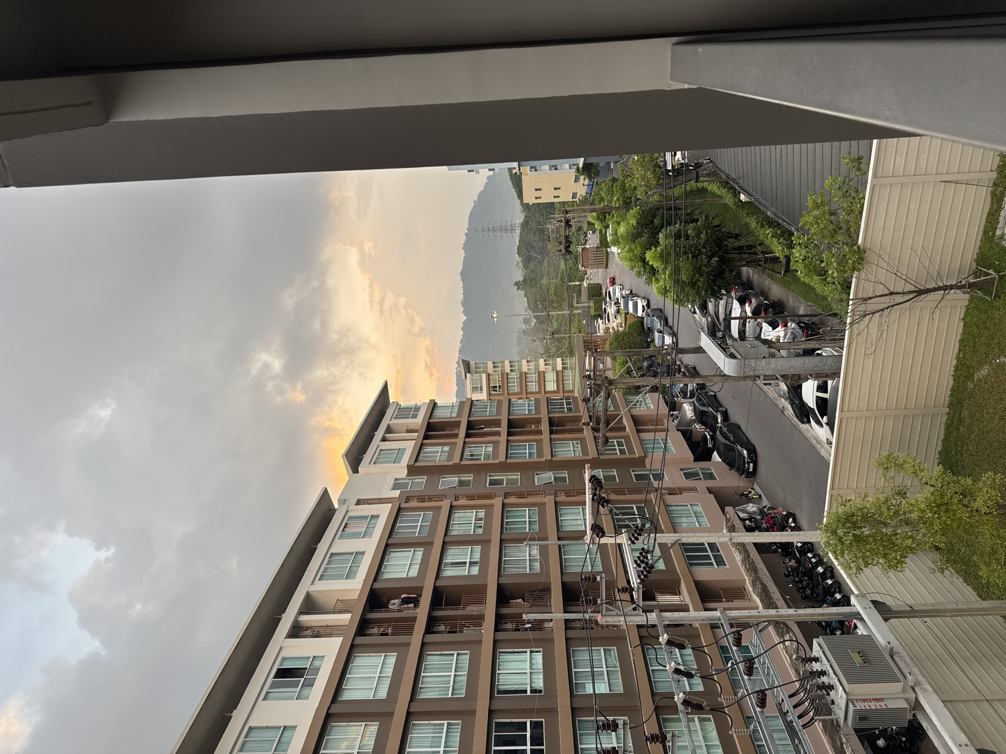 This image captures an urban scene in Kathu, Thailand, under a cloudy sky with hints of sunset. The foreground is dominated by the side of a modern, multi-story apartment building with brown and white facades, featuring numerous windows and balconies. Electrical infrastructure, including poles, wires, and a transformer box, is visible in front of the building. Below, a street runs alongside the building, lined with parked cars and motorcycles. Further down the street, more buildings and lush green trees are interspersed with the urban landscape. In the background, a mountain range is visible, silhouetted against the hazy sky, with a large electrical pylon standing out against the foliage. The overall impression is one of a densely populated residential area within a tropical environment, experiencing the transition from day to evening.