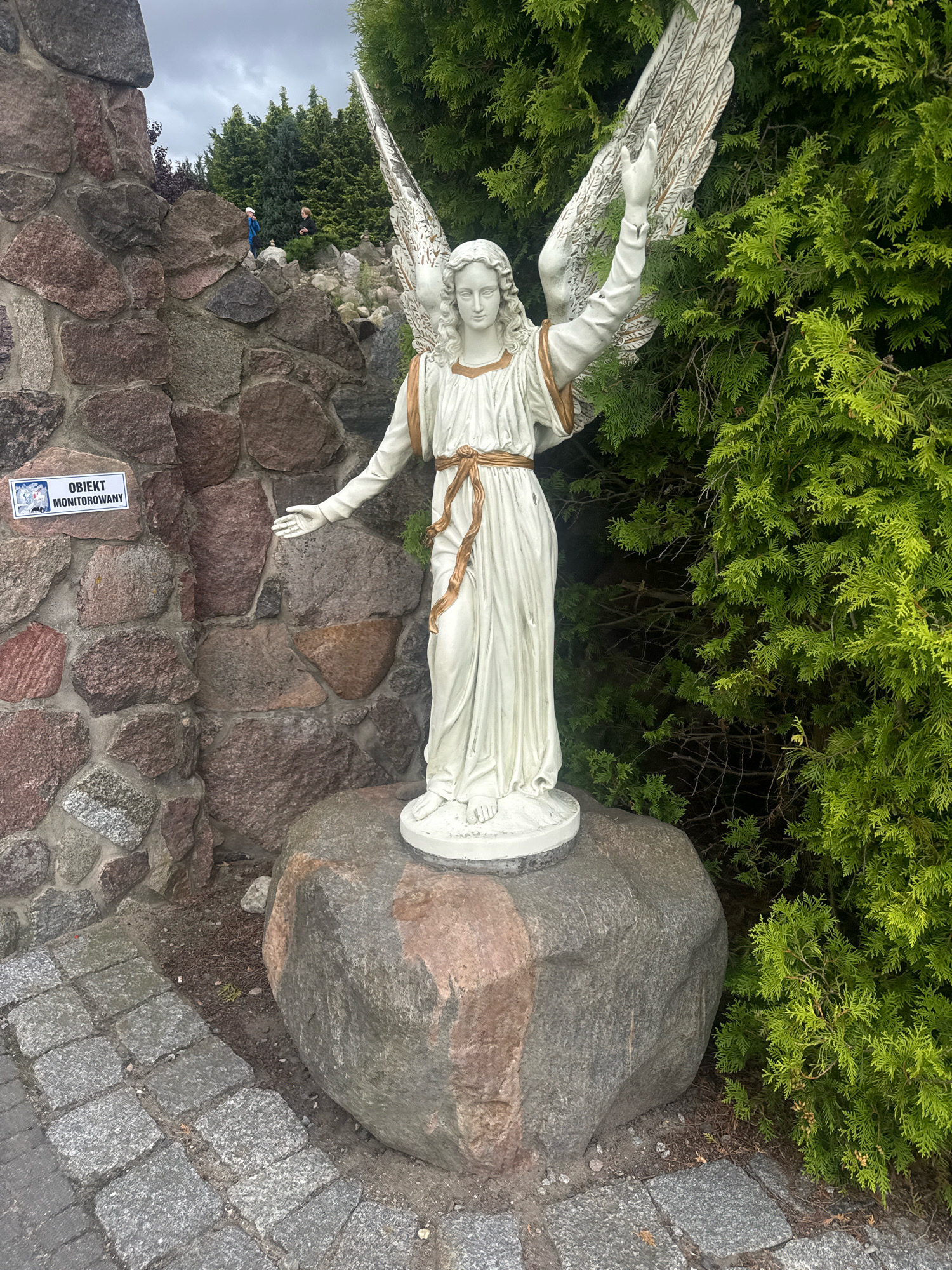 The image shows a white statue of an angel with outstretched arms and wings, standing on a large rock. The angel is dressed in a long, white robe with a gold sash. Behind the statue, there is a stone wall, a dense green hedge, and a grey, overcast sky. To the left of the statue, a sign attached to the stone wall reads "OBIEKT MONITOROWANY," which translates to "Monitored Object." In the background, on top of the stone wall, two small figures, possibly children, are visible among trees and rocks. The foreground shows a paved walkway made of grey cobblestones. This scene is located in Świebodzin, Poland, near the famous Christ the King statue.