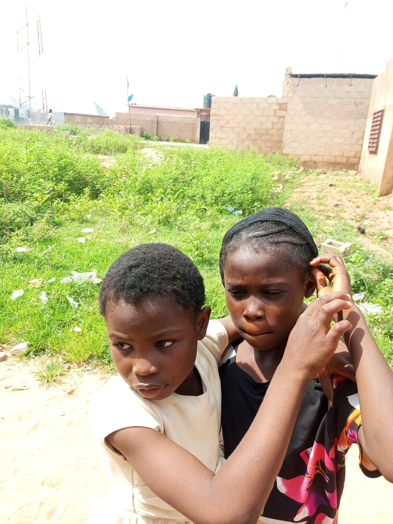 In the city of Tempure, Nigeria, two young girls are captured in a close-up outdoor portrait. The girl on the left, facing to her left, wears a light-colored garment and has short, dark hair. The girl on the right, facing forward with her head tilted slightly down, has her hair styled in cornrows. She is wearing a black top with a vibrant floral pattern of pink, yellow, and white flowers. Her right hand is raised to her ear, touching her hair. They are positioned against a backdrop of lush green foliage, with sandy ground in the foreground. In the background, a wall made of light-colored bricks is visible, along with parts of a building. Further in the distance, a tall communication tower with antennas stands against a bright, overcast sky.