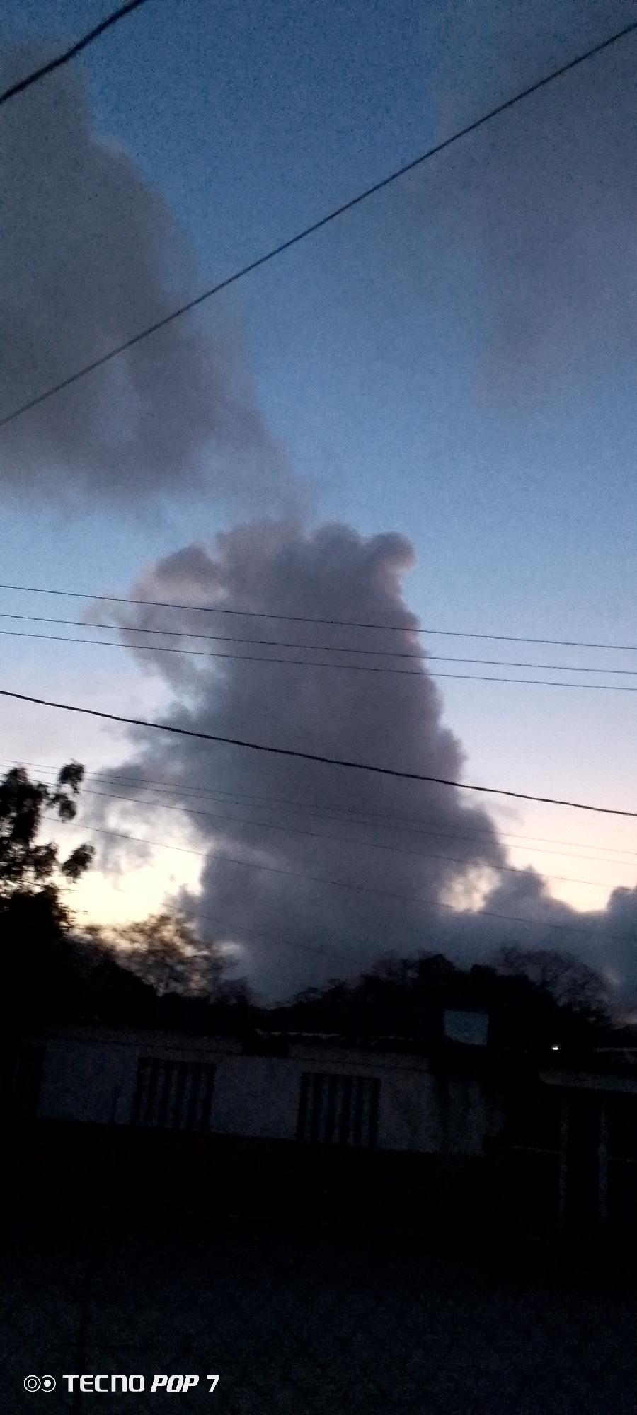 The image shows a cloudy evening sky over what appears to be a residential area in Altagracia, Venezuela. A prominent, dark cloud formation dominates the central sky, resembling a figure with a distinct head-like shape. Below the cloud, there are trees and the dark silhouette of a building with visible windows. Overhead, power lines cut across the sky. The lighting is dim, indicating either twilight or early evening. The lower portion of the scene is obscured by darkness. The bottom left corner shows the "TECNO POP 7" watermark, indicating the device used to take the photo.