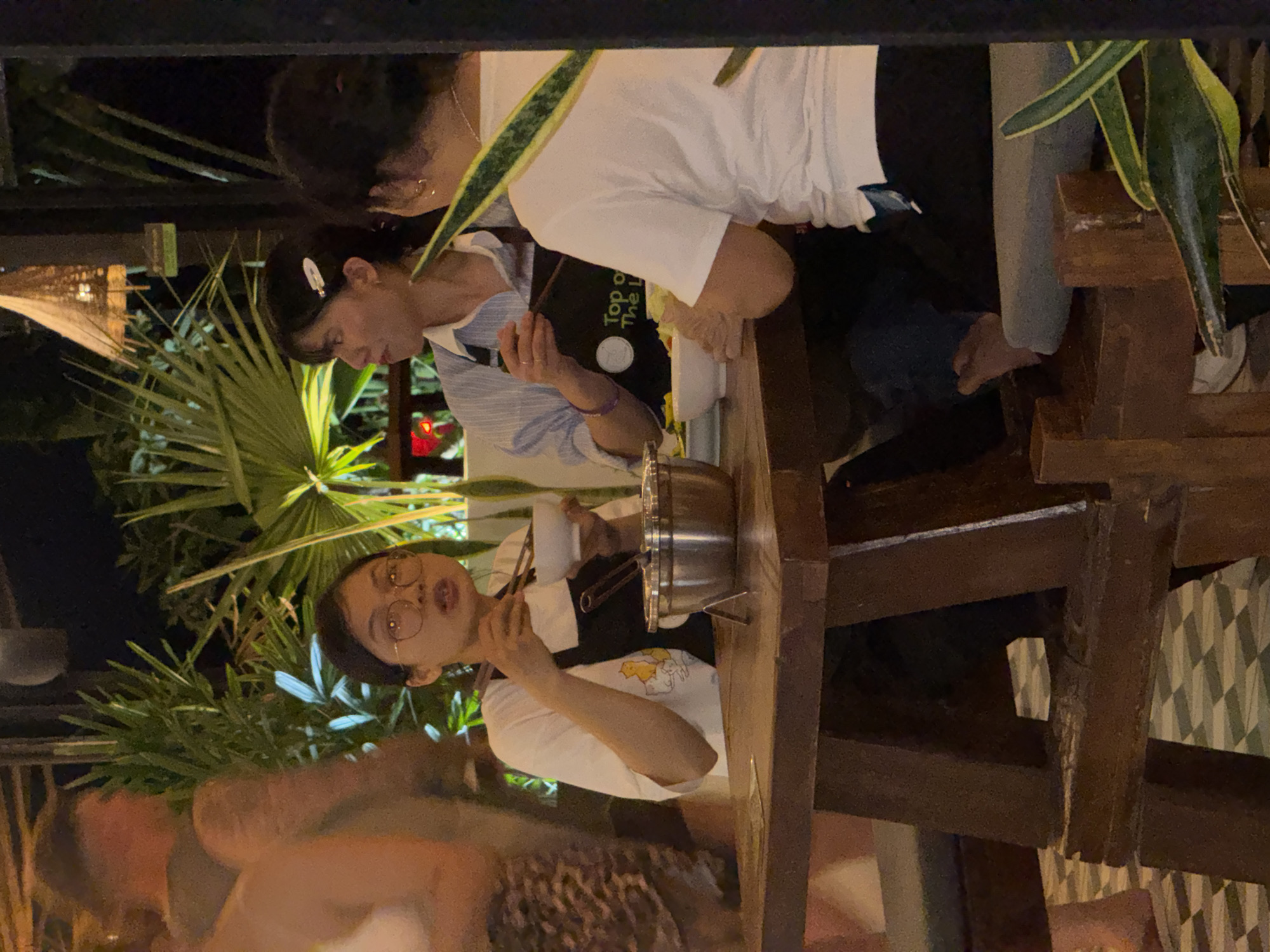 At night in Hà Giang 1 Ward, Vietnam, two women are sitting at a rustic wooden table outdoors, while another person stands partially visible behind them. One woman with short dark hair and glasses is eating from a stainless steel pot with chopsticks, wearing a white T-shirt under a dark apron. The other woman, with dark hair pulled back with a clip, is looking at a menu or booklet, wearing a light blue and white striped shirt. The environment includes lush green plants, suggesting a restaurant or outdoor dining space. The scene is framed by dark structural elements, possibly from the restaurant's architecture or overhead covering.