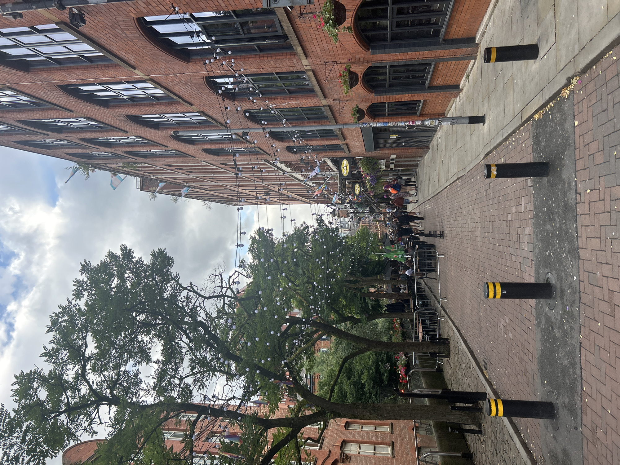 The image captures a street scene in the city of Manchester, United Kingdom, likely in the Northern Quarter, an area known for its Victorian architecture, independent businesses, and vibrant atmosphere. The perspective is from a street level looking down a cobbled lane lined with red brick buildings. On the right, a large brick building with multiple arched windows dominates the view, adorned with string lights and hanging flower baskets. On the left, more brick buildings are visible, interspersed with mature trees decorated with fairy lights and colorful flags. The street itself is paved with red bricks and features a sidewalk. Several bollards, painted black with yellow stripes, are positioned along the pedestrian area. A number of people are walking along the street, and outdoor seating areas for cafes or restaurants are set up along the left side. The sky is cloudy, suggesting an overcast day. The scene conveys a lively urban environment with a mix of architectural styles and a blend of commercial and pedestrian spaces.