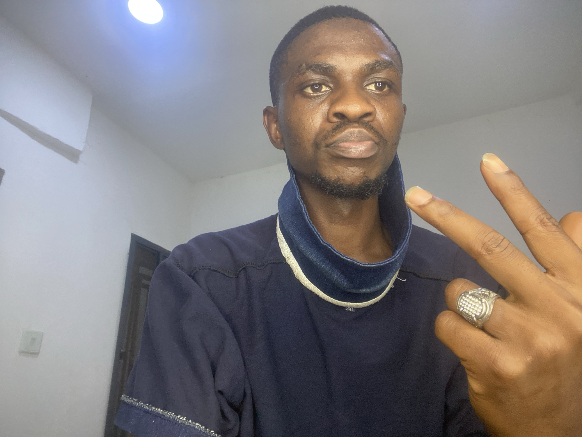 The image presents a chest-up portrait of a young Black man in an indoor setting. He has short dark hair, a trimmed beard, and a mustache, with dark eyes looking directly at the camera with a neutral expression. He is wearing a dark blue long-sleeved shirt featuring a distinctive high, stiff collar that appears to be denim with a white trim. The text "3XL" is visible on the collar area of his shirt. His right hand is raised, forming a "peace" or "victory" sign, and he wears a large, ornate silver-colored ring adorned with numerous small, sparkling stones on his ring finger.
The background consists of plain light-colored walls and a white ceiling. A bright, circular overhead light fixture provides illumination, and a white air conditioning unit is mounted on the wall to the left. A dark doorframe is partially visible on the right side of the frame, and a white rectangular object, possibly a light switch, is on the wall behind his right shoulder. The overall impression is a simple, well-lit indoor room. While the provided context suggests Abuja, Nigeria, there are no explicit visual cues within the image to confirm this location. The time of day is not discernible, as the primary light source is artificial.