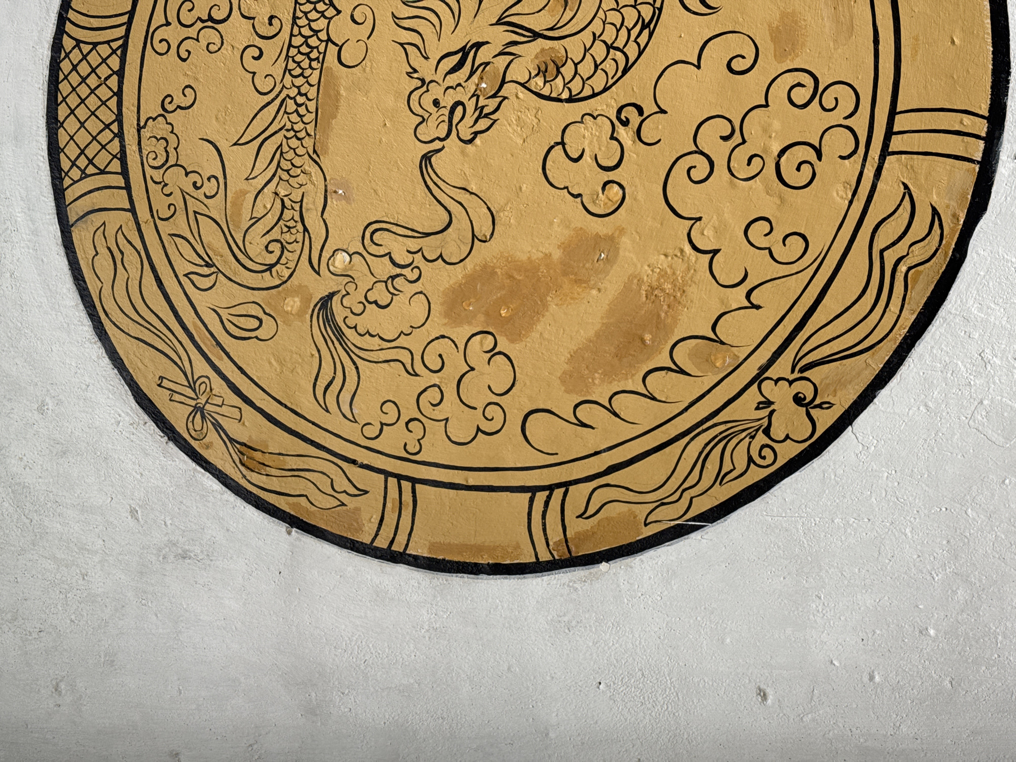 A partial view of a circular mural is painted on a white wall. The upper half of the mural shows a golden dragon surrounded by stylized clouds and traditional patterns, all outlined in black. The wall's texture is rough. This is a wall in Sà Phìn Commune, Vietnam.