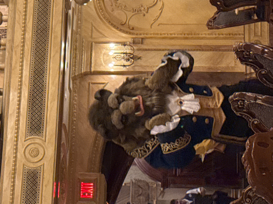 This image depicts an indoor scene within the Be Our Guest Restaurant, located in Magic Kingdom at Walt Disney World Resort, Bay Lake, Florida, United States.
A costumed performer portraying the Beast character from Disney's "Beauty and the Beast" stands centrally. The performer wears a brown fur costume featuring a pronounced snout, fangs, and an open mouth with visible pink gums. The costume includes a dark blue tailcoat with elaborate gold embroidery on the lapels and cuffs, a tan-colored waistcoat, a white ruffled jabot, and white ruffled cuffs. The performer's left arm is slightly bent, with the hand raised to chest height.
The background features opulent architecture consistent with a grand dining hall. Walls are a light golden-brown, adorned with intricate decorative moldings, fluted pilasters, and stylized bas-relief patterns. A dark metal wall sconce with three illuminated candle-like bulbs is mounted on the wall to the right. In the upper left, a red "EXIT" sign with white lettering is illuminated above a dark archway. Partially visible in the lower left background, a male individual wearing a white shirt and dark vest suggests a staff member. The immediate foreground shows the dark, ornate backs of several dining chairs, indicating the presence of tables. The scene is illuminated by ambient artificial light.