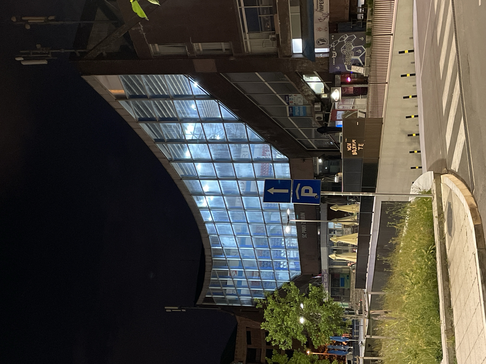 This is an outdoor shot, likely at night, showcasing a street scene in Zemun Urban Municipality, Serbia. A pedestrian walkway or bridge covered by a glass-paneled arch is a prominent feature of the scene. A blue parking sign with an arrow pointing left is visible in front of the glass structure. To the right of the archway is a building with businesses. There are parasols outside of an establishment. Below that building, some greenery and fencing are noted. The sidewalk and the street can be seen on the right of the image. There is also a crosswalk striped in black and white.