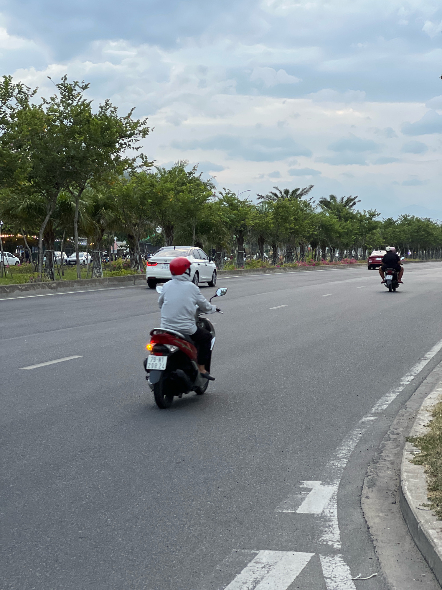 The image shows a street scene in Nha Trang, Vietnam, likely during overcast weather. In the foreground, a person wearing a light gray long-sleeved shirt and a red helmet rides a red and black scooter. The scooter has a license plate that reads "79-M1 298.24". The scooter's rear brake light is illuminated. Further down the road, another scooter with two passengers, one wearing a pink helmet, is visible. In the background, several white cars are parked and driving on the road, including a white Honda sedan. The street is lined with numerous trees, including palm trees, and a cloudy sky dominates the upper portion of the image. The road has white lane markings and a concrete curb on the right side, with dry grass and weeds growing along the edge.