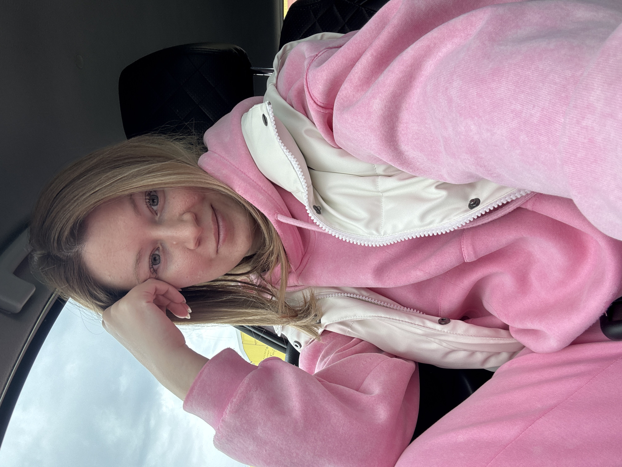 A woman with blond hair is seated in a car, resting her face on her hand. She is wearing a pink hoodie and a white vest. The car's interior is visible, including the headrest and dashboard. The sky is overcast and visible through the car window. The location is likely in a vehicle in the city of Chaikovsky, Russia.
