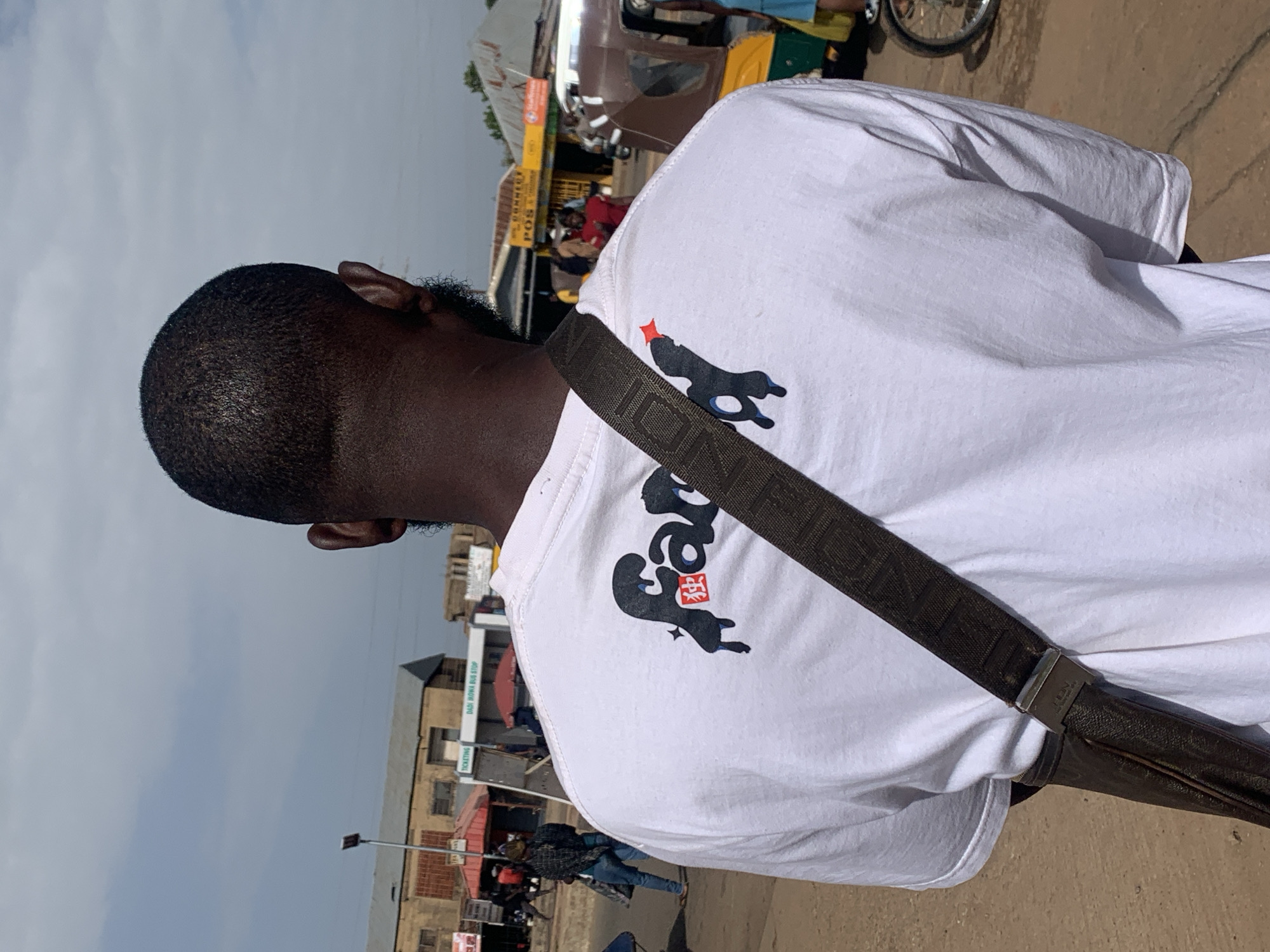 In Gura Top, Nigeria, a Black male is captured from behind, wearing a white t-shirt with a black graphic design and a dark brown crossbody bag. The graphic on the t-shirt features stylized text and Asian characters. The man stands on a dirt road, with buildings and a bus stop visible in the background. A rickshaw and other people are present in the bustling street scene. The sky is partly cloudy.