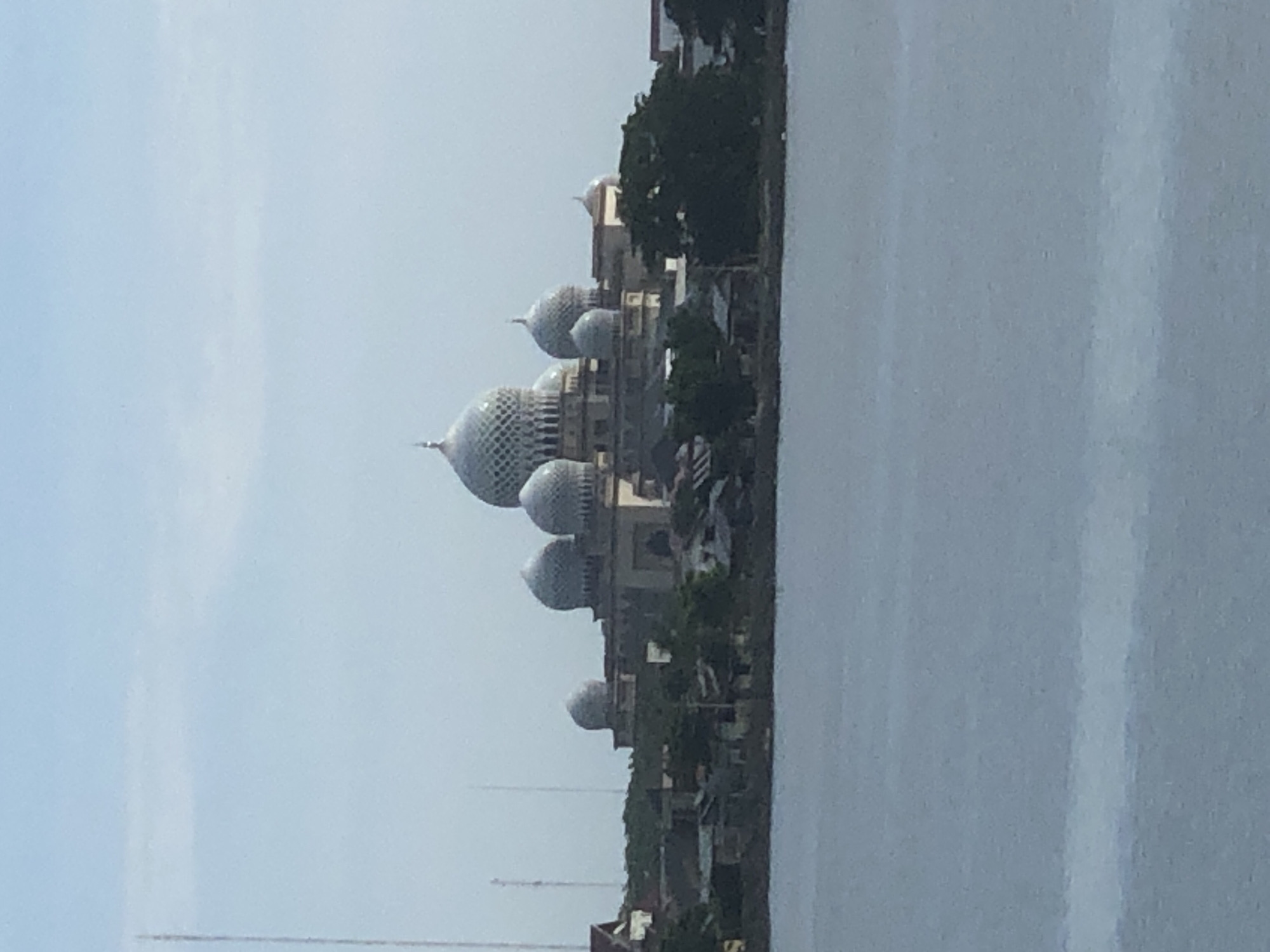 The image shows Baiturrahman Mosque in Lhokseumawe, Indonesia, situated along a body of water. The mosque is recognizable by its large central dome and several smaller surrounding domes, all featuring a unique lattice pattern. The mosque complex is surrounded by trees and buildings. The sky is overcast.
