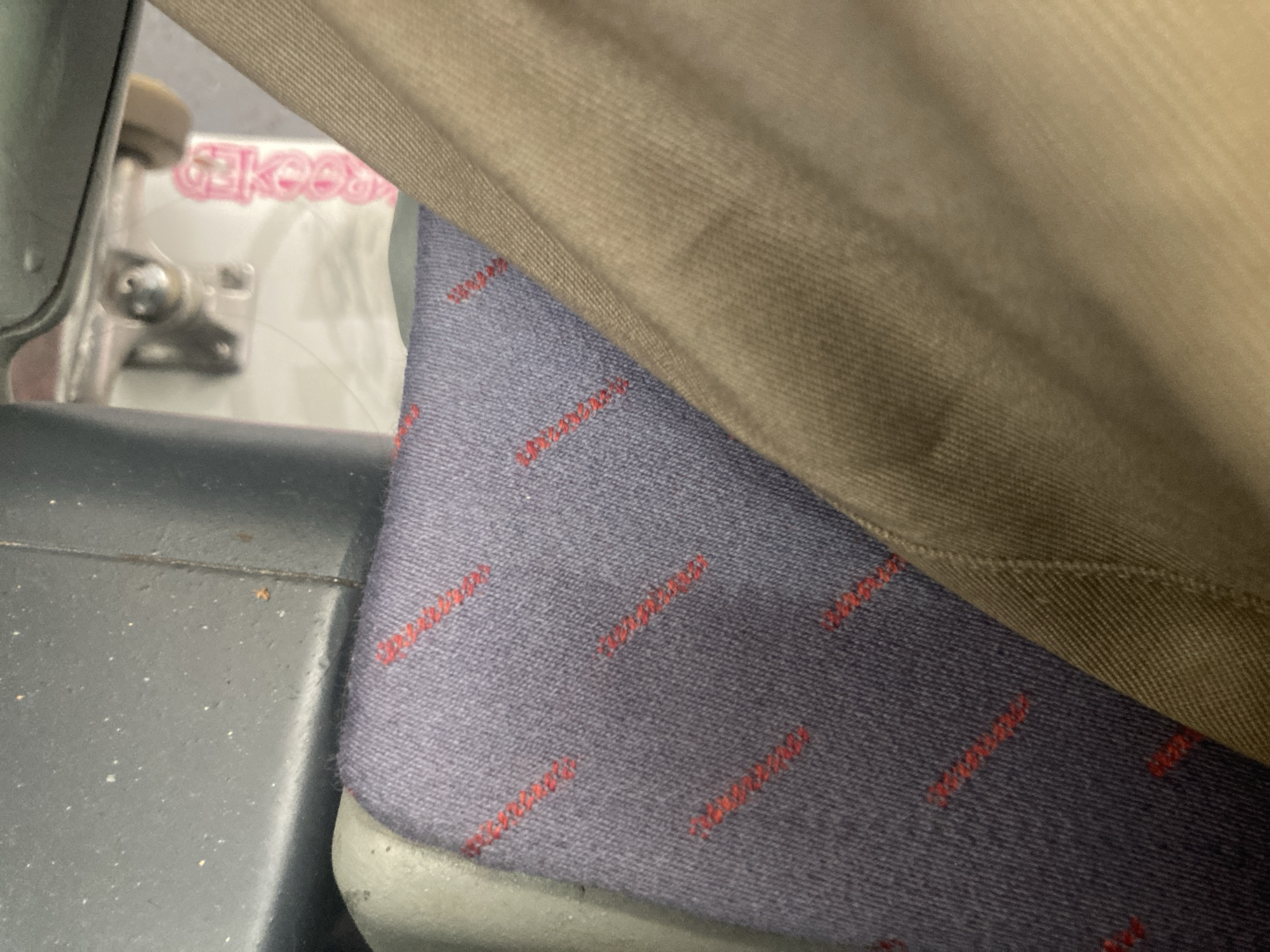 The image shows a close-up view of a seat on public transport. The seat is upholstered in a dark blue fabric with a repeating pattern of thin red lines. A person's light brown pants are visible in the foreground, partially obscuring the seat. In the background, through a gap in the seat structure, a white package with pink text is visible, possibly the packaging for a skateboard. The overall impression is one of a utilitarian interior, likely a bus or train. The image does not contain any visual information that would allow for the determination of its location in Montcada i Reixac, Spain.