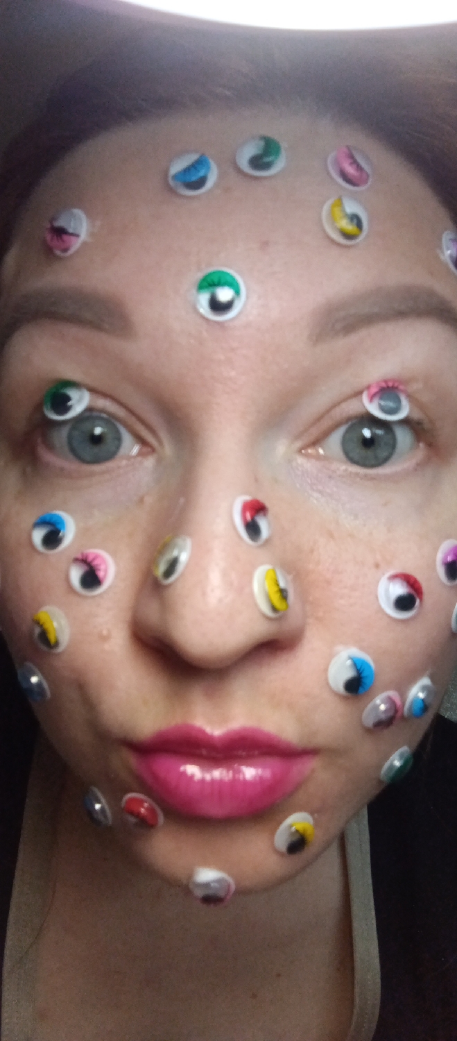 A close-up, slightly off-center portrait features a person, likely female, shown from the shoulders up, facing the camera directly. Their face is adorned with numerous small, multi-colored googly eyes. These plastic eyes, with movable pupils, are adhered across the forehead, around the natural eyes, on the bridge and sides of the nose, on the cheeks, and scattered across the chin and jawline. The googly eyes display a variety of colors including blue, green, pink, yellow, red, and silver/grey, and vary slightly in size.
The person's natural grey-blue eyes are clearly visible, looking straight ahead, with several googly eyes placed strategically around them, including one almost on the eyelid of the right eye. Their eyebrows are well-defined. The lips are painted with a glossy, vibrant pink lipstick and are slightly pursed. The overall presentation suggests a playful, artistic, or costume-related activity. The background is indistinct, and the lighting indicates an indoor setting. No text is visible in the image.