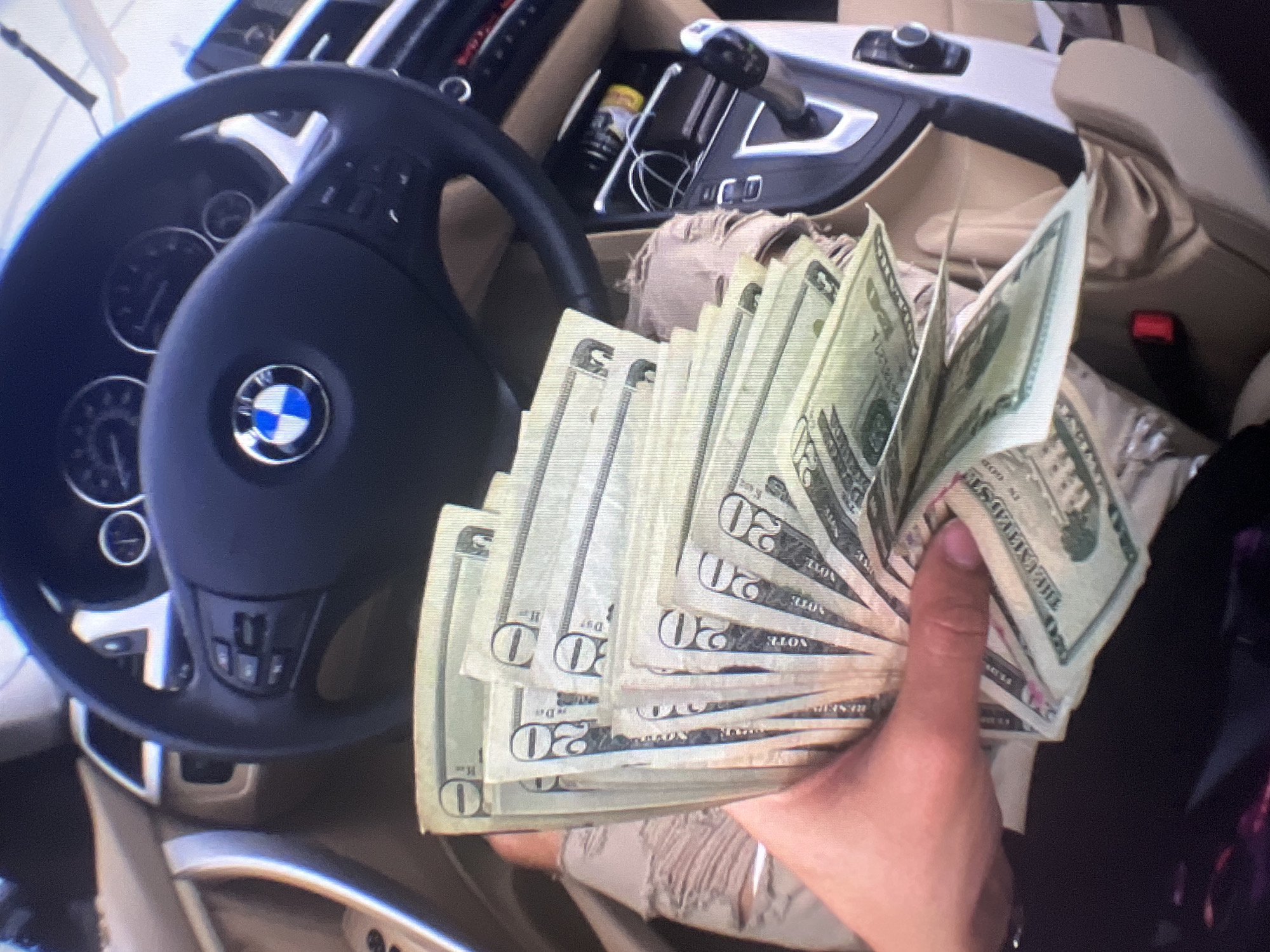 The image displays the interior of a car, specifically the driver's side, situated in Varna, Bulgaria. The primary subject is a person's hand, positioned in the lower right, holding a large fan of United States twenty-dollar bills.
The car's interior features a black steering wheel with a prominent BMW logo, indicating the vehicle's make. Parts of the dashboard and instrument cluster, including circular gauges, are visible behind the steering wheel. To the right, the center console is equipped with a gear shifter and control buttons. A cup holder containing what appears to be a Red Bull can and a charging cable is also seen. The car's upholstery is a light beige or tan color, and a red seatbelt buckle is partially visible in the background.
The banknotes are clearly identifiable as US twenty-dollar bills, with the number "20" and the text "TWENTY DOLLARS" visible on several notes. Text such as "THE UNITED STATES" can also be discerned on some of the bills. The money is fanned out, suggesting a deliberate display.
The scene captures a moment of presenting a substantial amount of cash within a vehicle's setting. The lighting suggests an indoor or shaded environment, typical for a car interior, without specific indications of time of day or weather.