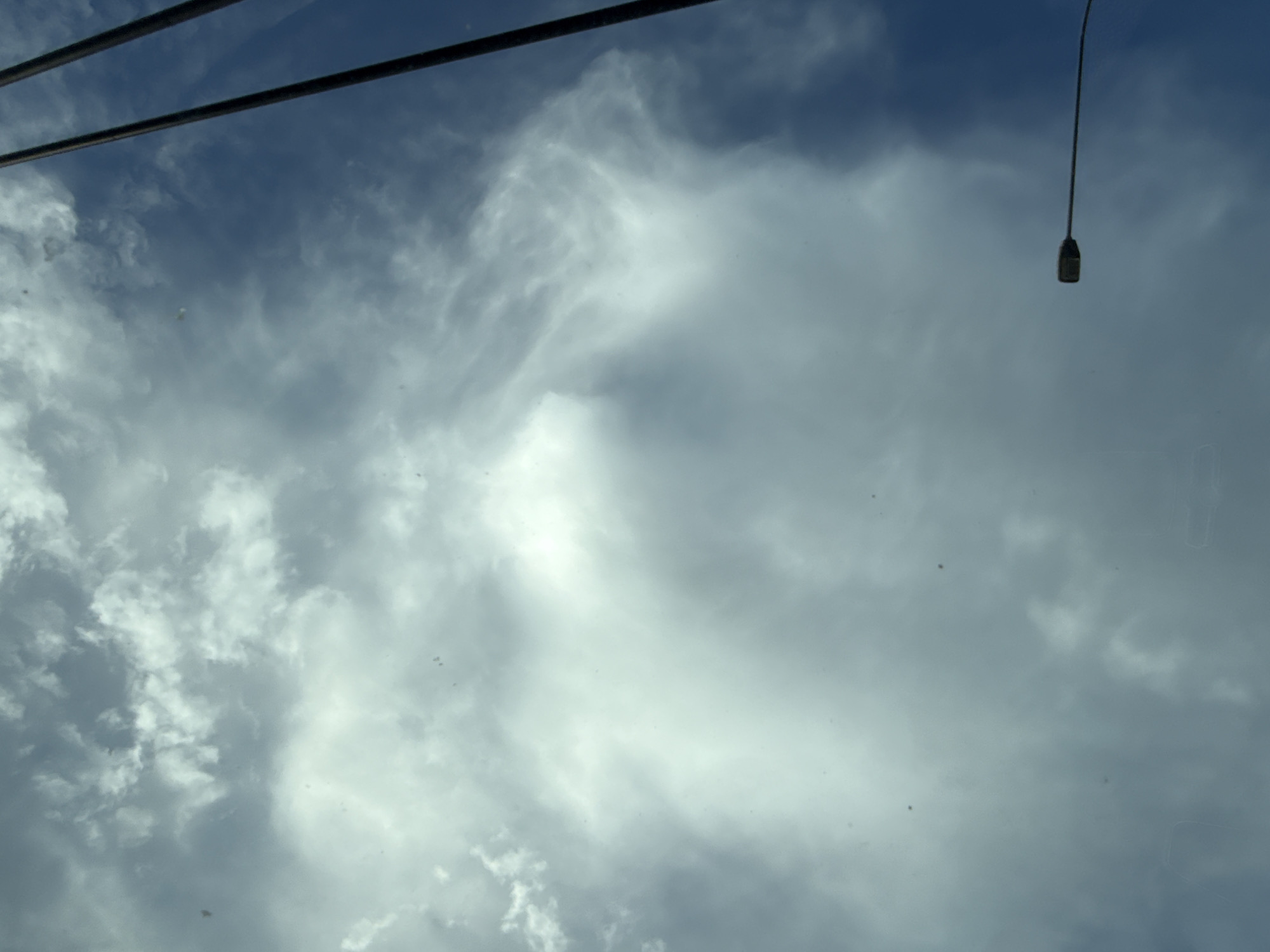 A low-angle shot captures a cloudy sky with a visible streetlamp in the foreground. The sky is a mix of dark blue and white clouds, with some areas appearing brighter than others. The streetlamp is dark and elongated, with a rectangular light fixture. Two dark vertical lines, possibly wires or poles, are visible on the right side of the image, extending towards the top. There are several small, dark specks scattered across the sky, which may be dust or debris on the lens or camera. The image appears to be taken during the daytime, with the lighting suggesting overcast conditions. The scene is devoid of any people or specific urban landmarks that would immediately indicate its location within Wrocław, Poland.