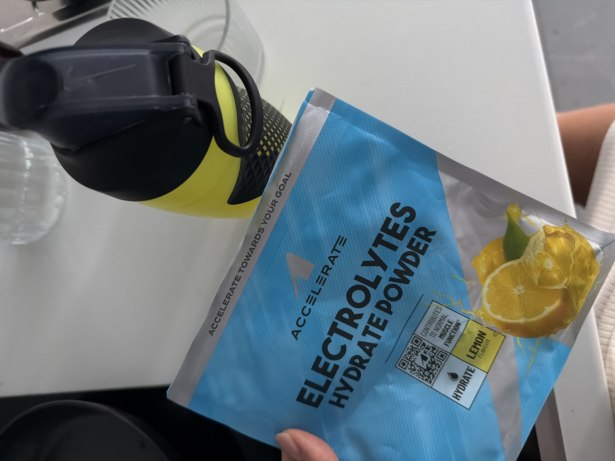 The image shows a close-up of a blue and silver packet of "Electrolyte Hydrate Powder" with a lemon flavor. The packet is partially held by a person's hand. The powder packaging features text like "ACCELERATE TOWARDS YOUR GOAL" and "CONTRIBUTES TO NORMAL MUSCLE FUNCTION". A QR code is visible on the packet. To the right of the packet, a bright yellow water bottle with a black lid and handle is partially visible. The objects are placed on a white surface, likely a countertop, with a portion of a dark pan visible in the upper left corner. The background is out of focus, but appears to be an indoor setting. The scene is lit by artificial light. The specific location in Finestrat, Spain is not determinable from the image.