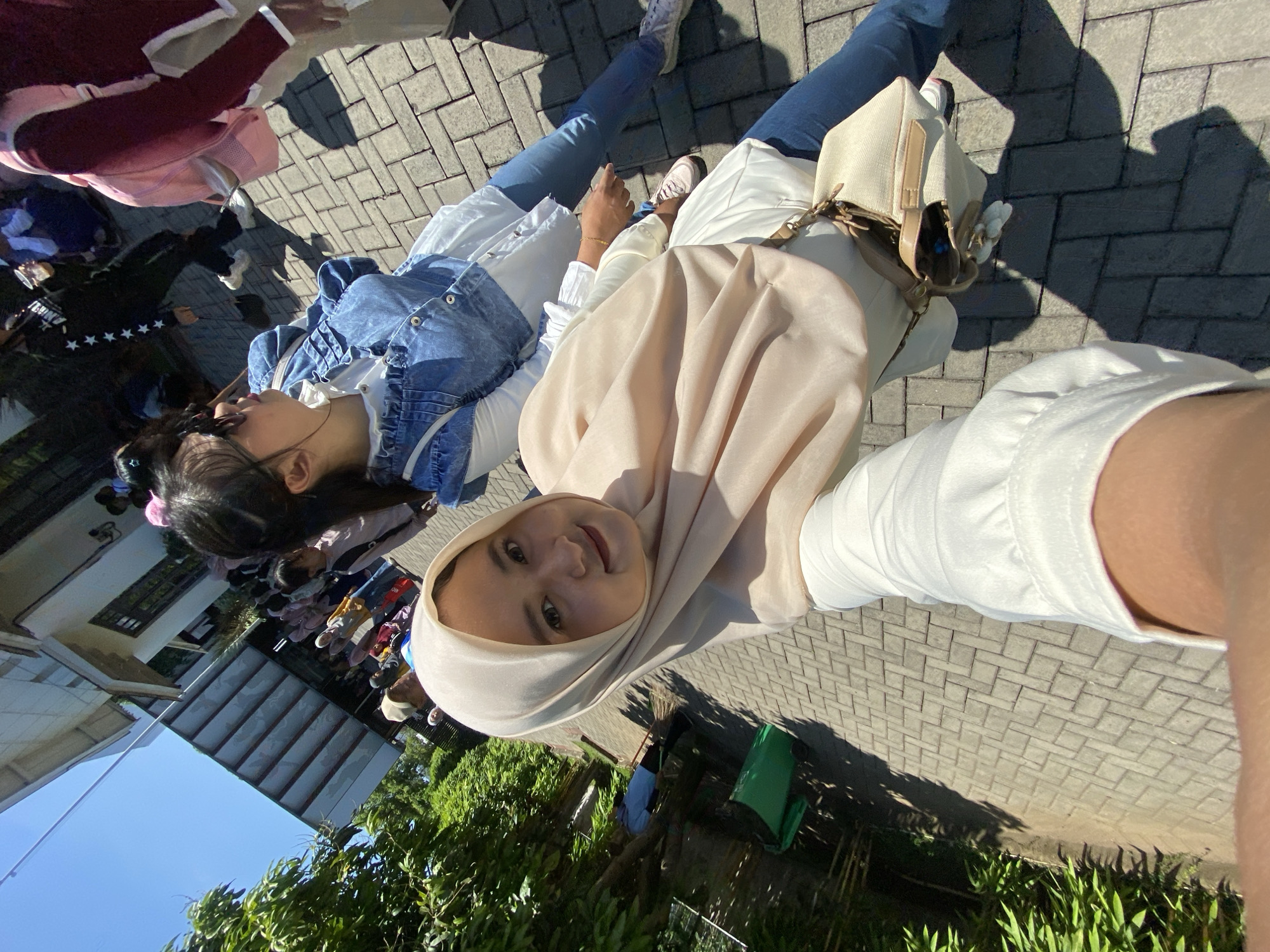 A high-angle selfie shows two women in Mangli, Indonesia. The woman in the foreground wears a beige hijab and a white top. Her companion is dressed in a white top with a denim jacket. Both women wear jeans and sneakers. A beige handbag is visible near the hijab-wearing woman. In the background, other people, including school children, are visible near a building. A blue sky and green foliage are also visible. The ground is paved with interlocking grey bricks.