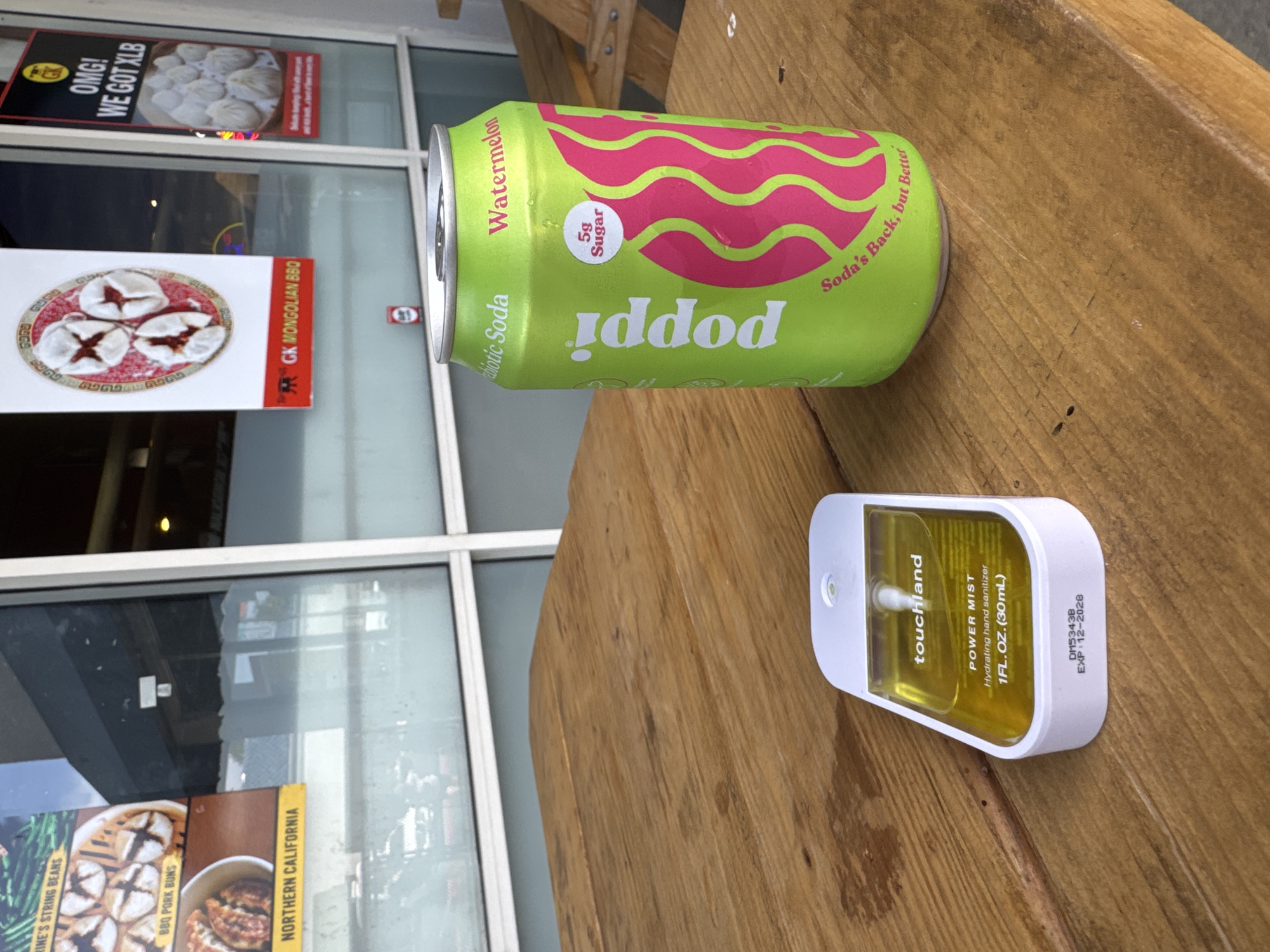 A close-up, eye-level shot captures a wooden picnic table outdoors, with a bright green can of Poppi Watermelon Probiotic Soda and a white and yellow Touchland Power Mist hydrating hand sanitizer prominently displayed. The soda can, positioned to the right, features a vibrant pink wave design and text indicating "Poppi," "Watermelon," and "5g Sugar." To the left, the hand sanitizer is clear with a yellow liquid, a white sprayer, and text indicating "Touchland," "POWER MIST," and "1FL.OZ. (30mL)."
In the background, seen through a large glass window, are posters advertising food items. One poster on the left advertises "VEGETABLE'S STRING BEANS" and "BBQ PORK BUNS" with "NORTHERN CALIFORNIA" at the bottom. Another poster to the right advertises "OMG! WE GOT XLB" with an image of dumplings. Below these posters, the text "GK MONGOLIAN BBQ" is visible. The setting appears to be an outdoor dining area of a restaurant. The image is likely taken in Roseville, California, United States of America, given the restaurant advertisements that suggest Asian cuisine and potentially a regional focus. No people are visible in the frame.