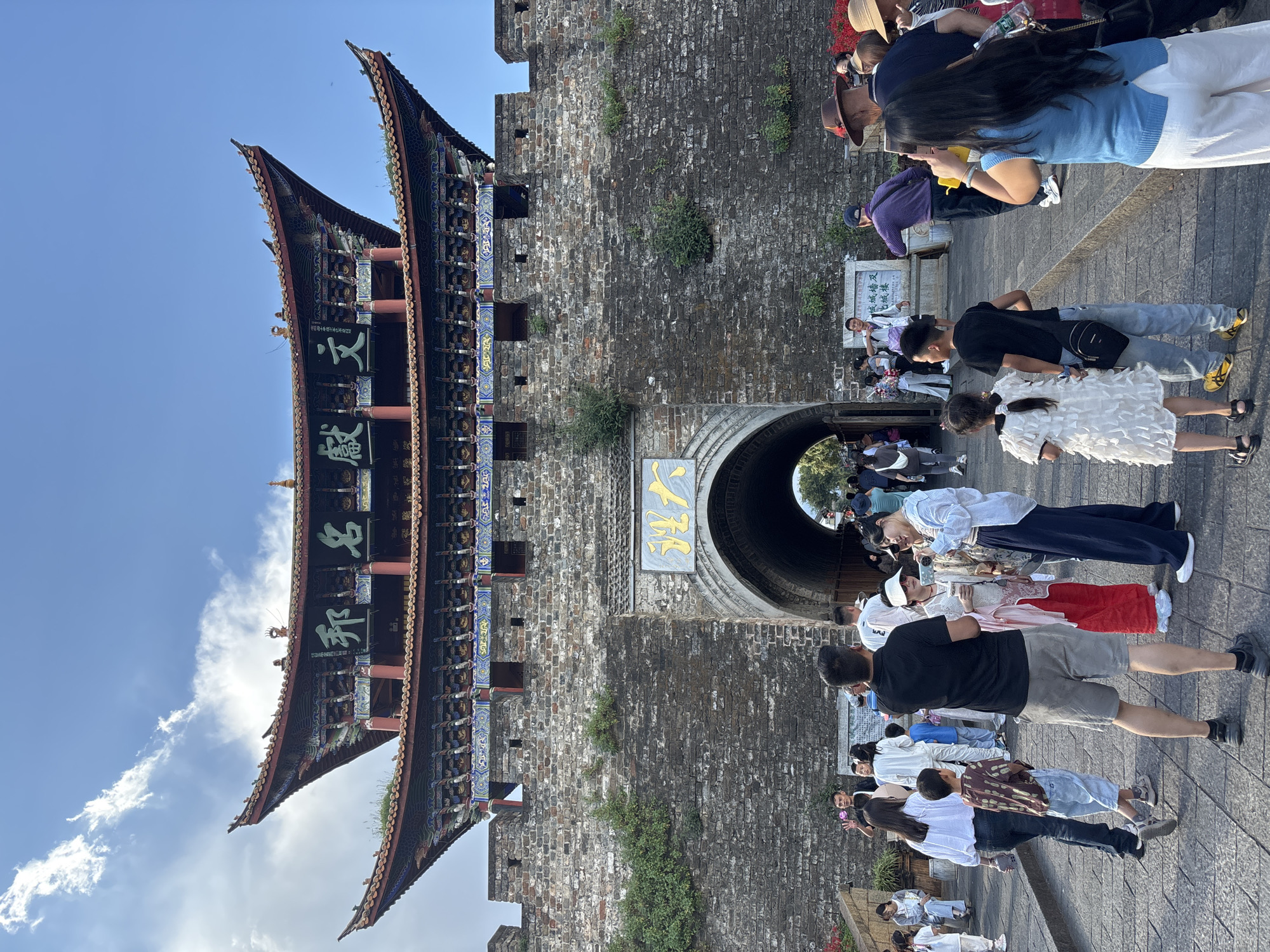 This is a full, eye-level shot of the Xi'an Gate in Dali City, China. The gate is a large, ancient structure made of stone and wood. It is topped with a traditional Chinese roof with upturned eaves, decorated with colorful carvings and Chinese characters. The gate has a large archway through which people are entering and exiting. The wall surrounding the gate is made of weathered stone blocks. In front of the gate, a crowd of people, dressed in casual, modern clothing, are gathered. Some are taking pictures with their phones, while others are walking and talking. The ground is paved with cobblestones. The sky is blue with a few scattered white clouds. The overall scene depicts a popular tourist destination with a blend of historical architecture and contemporary human activity.
