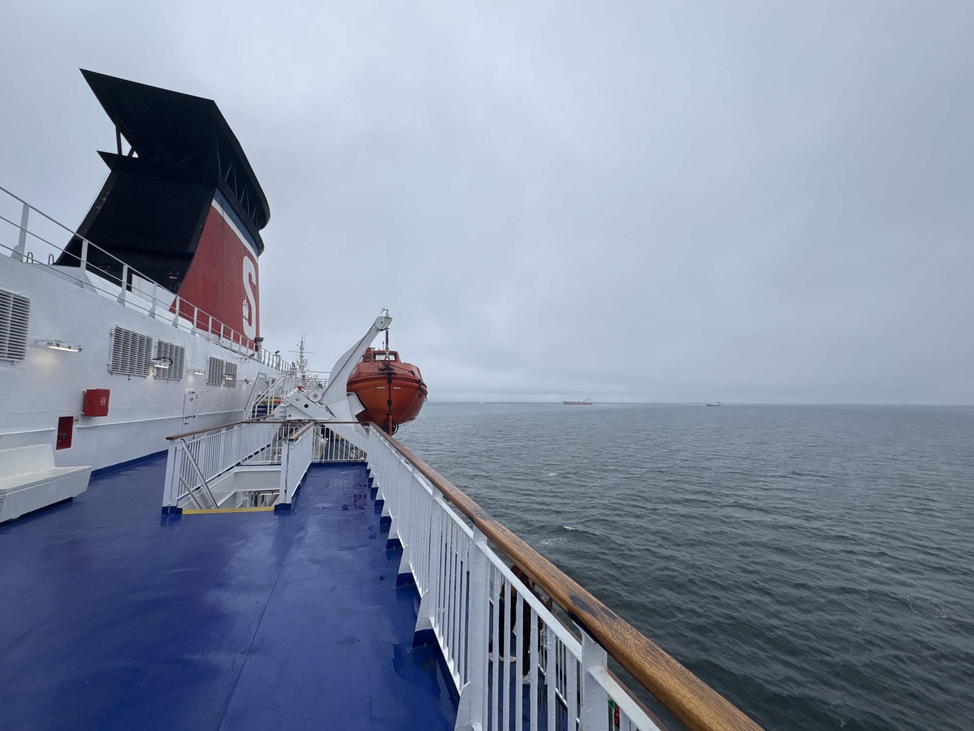 The image depicts the deck of a large ferry, likely the Stena Line ferry operating between Gdynia, Poland, and Karlskrona, Sweden. The perspective is from the upper deck, looking outwards towards the Baltic Sea under a cloudy sky. The ferry's distinctive red and white superstructure with a large black funnel is visible on the left. A bright orange lifeboat is mounted on the side of the vessel. The deck itself is painted a vivid blue, with white railings and a wooden handrail. The sea is choppy, indicating a moderate swell. In the distance, several cargo ships are visible on the horizon. The overall atmosphere is subdued, consistent with overcast weather conditions.