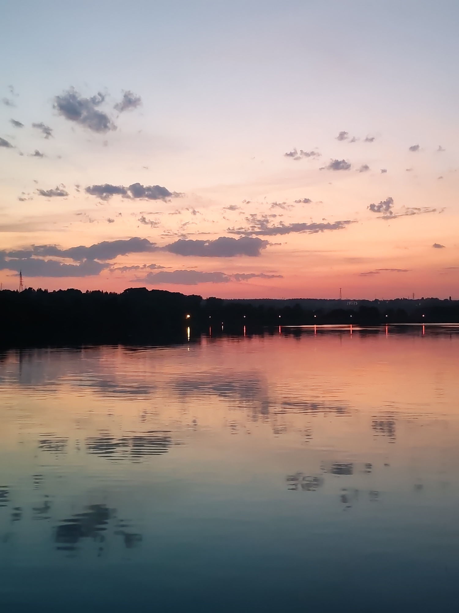 A wide-angle shot captures a tranquil body of water reflecting a sunset or sunrise sky. The sky is gradient, transitioning from light blue and pale peach at the top to orange-pink near the horizon. Scattered cumulus clouds with grey underbellies are visible in the sky. A dark, silhouetted treeline forms the horizon, with sparse light fixtures punctuating its length. These lights, along with reflections of clouds and the gradient sky, are mirrored on the water's surface, creating a textured effect. The water deepens in color from the horizon to the foreground, starting with orange and pink reflections near the horizon and shifting to a blend of blue and teal in the foreground. There are no people visible. The scene appears serene and still. The location of Ermolino, Russia is presumed based on an external image caption. The presence of possible industrial structures on the horizon suggest a suburban/rural environment.
