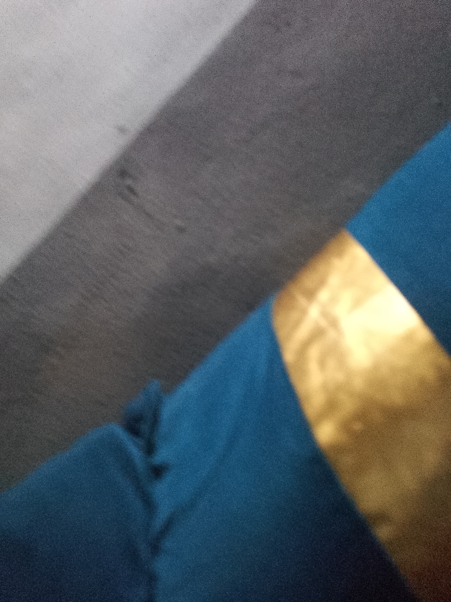 An extreme close-up, abstract image shows a textured, grey surface divided by a diagonal shadow into a lighter and darker area. A band of bright, metallic gold runs diagonally across the image, partially obscuring the grey surface and casting a shadow of its own. Below the gold band, a swath of dark teal fabric is visible, featuring subtle folds and creases. The overall impression is of a highly textured, abstract composition with a strong diagonal emphasis. There is no discernible information about Jauro Yinu, Nigeria.