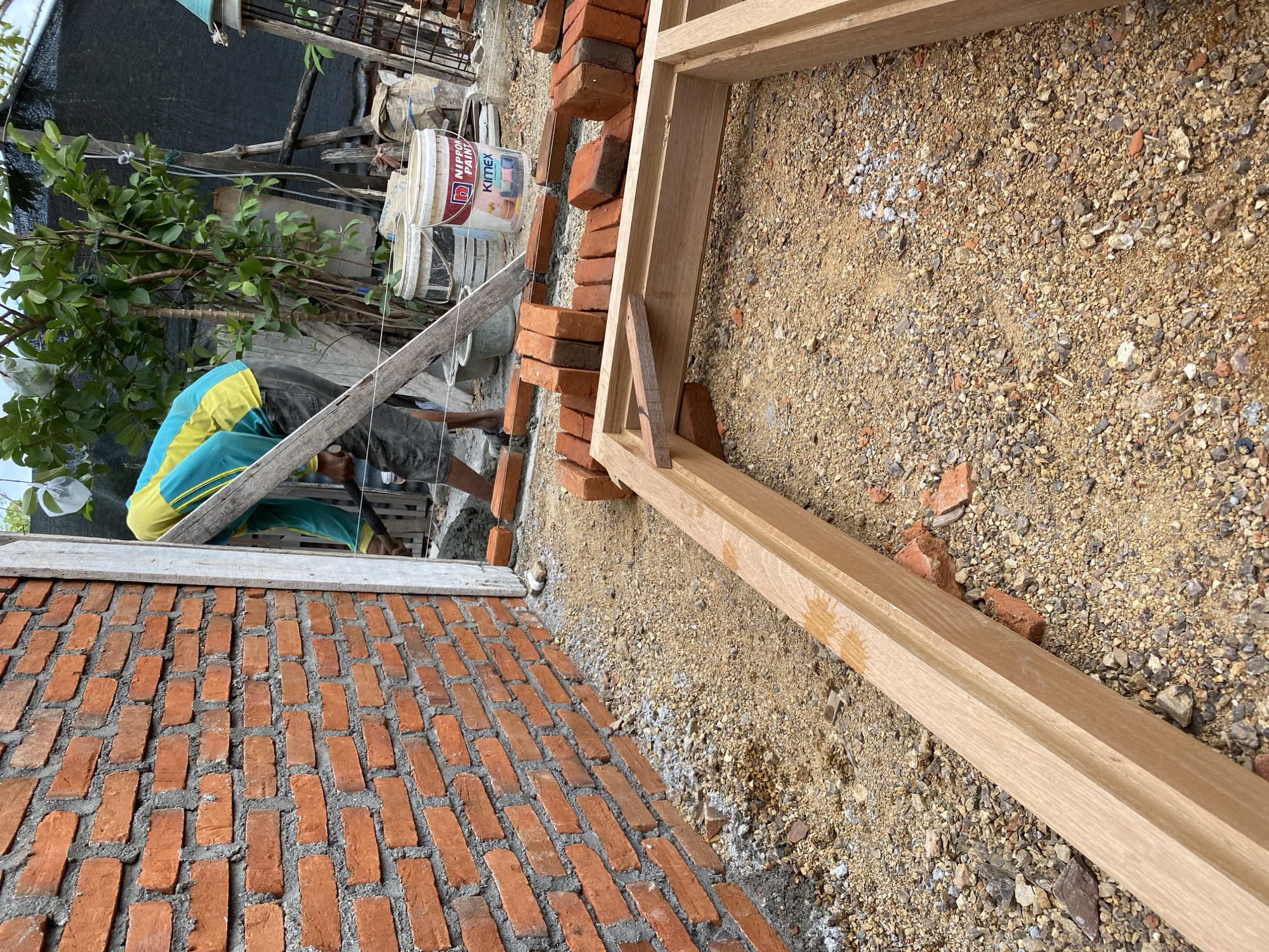 The image depicts a construction site, likely in Banda Aceh, Indonesia, where a brick wall is being built. A construction worker, wearing a yellow and teal long-sleeved shirt and dark shorts, is actively engaged in the building process. He is positioned next to the unfinished brick wall, holding a tool, likely for spreading mortar or placing bricks. The ground is covered with loose soil, gravel, and scattered bricks, indicating ongoing construction work. A wooden door frame lies in the foreground, suggesting it is either about to be installed or has been prepared for the opening. Several buckets, including one labeled "NIPPON PAINT KIMEX," are visible in the background, along with building materials and vegetation. The overall scene portrays a typical outdoor construction environment.