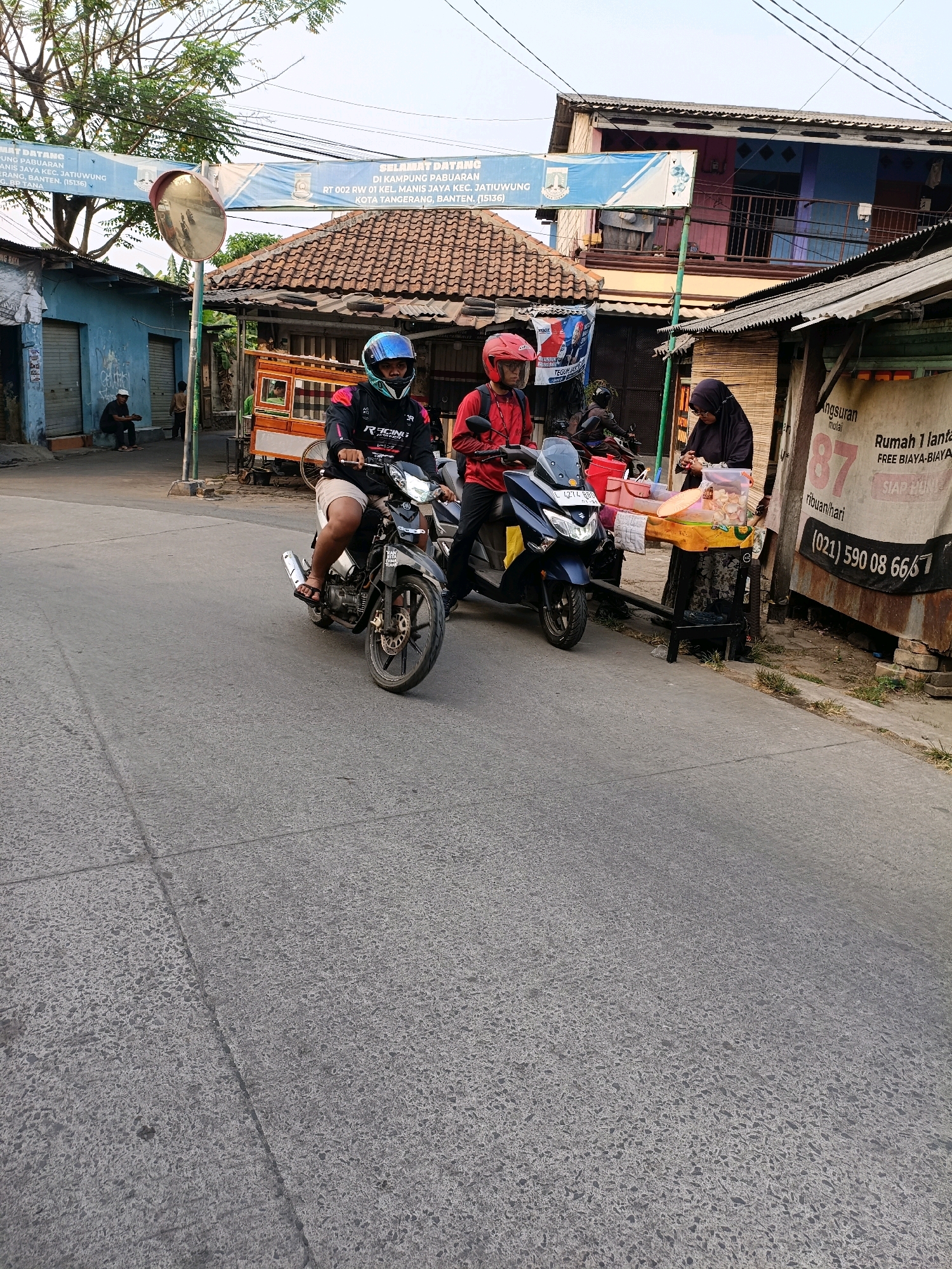 In Tangerang, Indonesia, a street scene unfolds with several people and vehicles. Two individuals are seated on a sidewalk on the left, partially obscured by a blue building with closed shutters. In the foreground, two men ride motorcycles. The man on the left rides a black motorcycle, wearing a black and red jacket and a blue helmet. The man on the right rides a blue scooter, wearing a red jacket and a red helmet. A street vendor, a woman wearing a black hijab, stands behind a small stall, arranging food items. The stall is adorned with a banner advertising "Rumah 1 lantai" (1-story house) with the text "FREE BIAYA-BIAYA" (free of charges) and a phone number "(021) 590 08 66/7". A large banner overhead displays "SELAMAT DATANG DI KAMPUNG PABUARAN" (Welcome to Paburan Village). The environment suggests a bustling urban setting with low-rise buildings and a paved road.