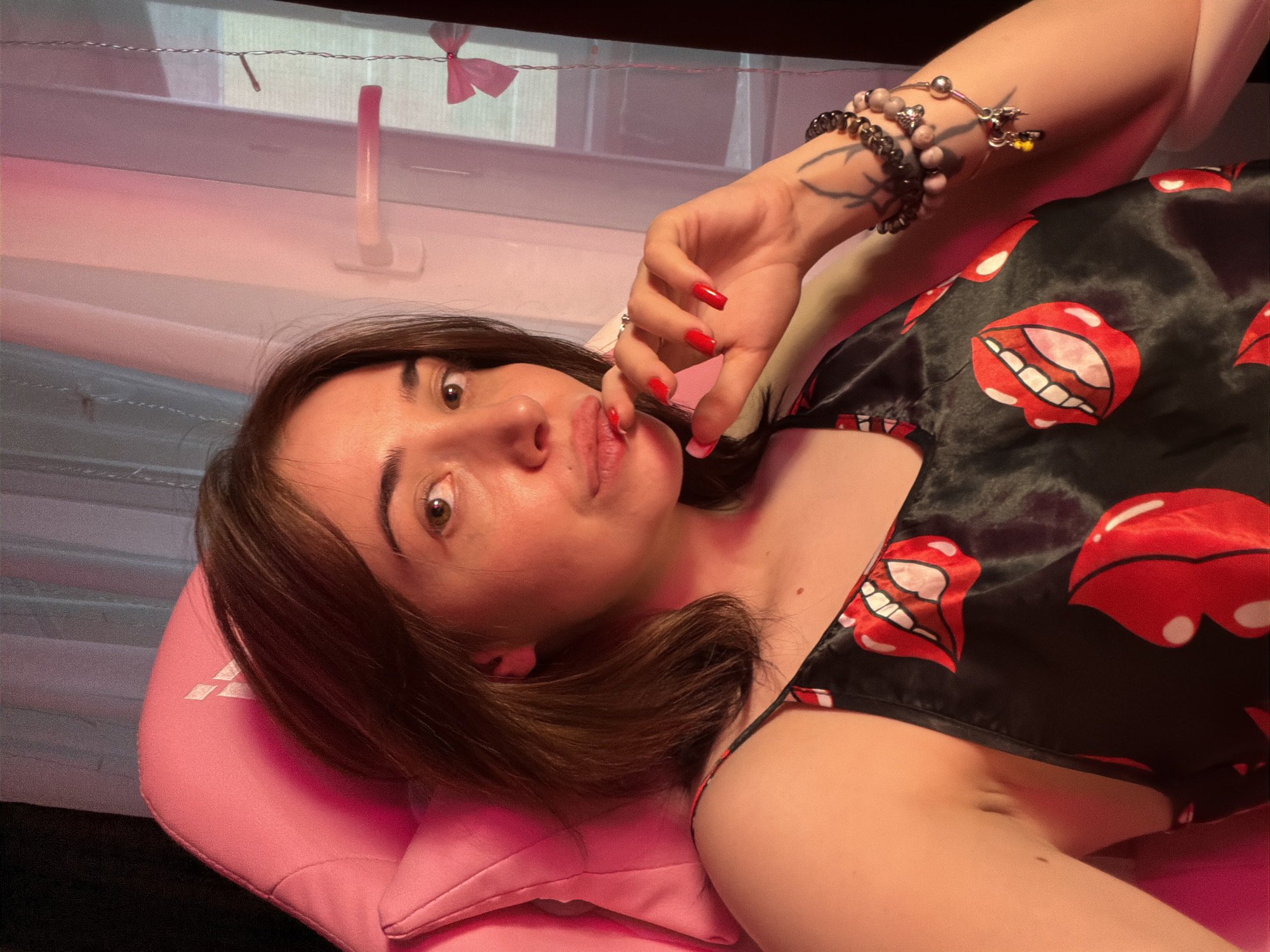 A young woman with medium-length brown hair and brown eyes is captured in a selfie. She is seated in a pink gaming chair, her right hand touching her lips. She is wearing a black camisole with a red lip print pattern. On her left wrist, she has several bracelets, and a tattoo is visible on her forearm. The background shows a window with sheer pink curtains and a light pink interior. The lighting in the scene is predominantly pink.