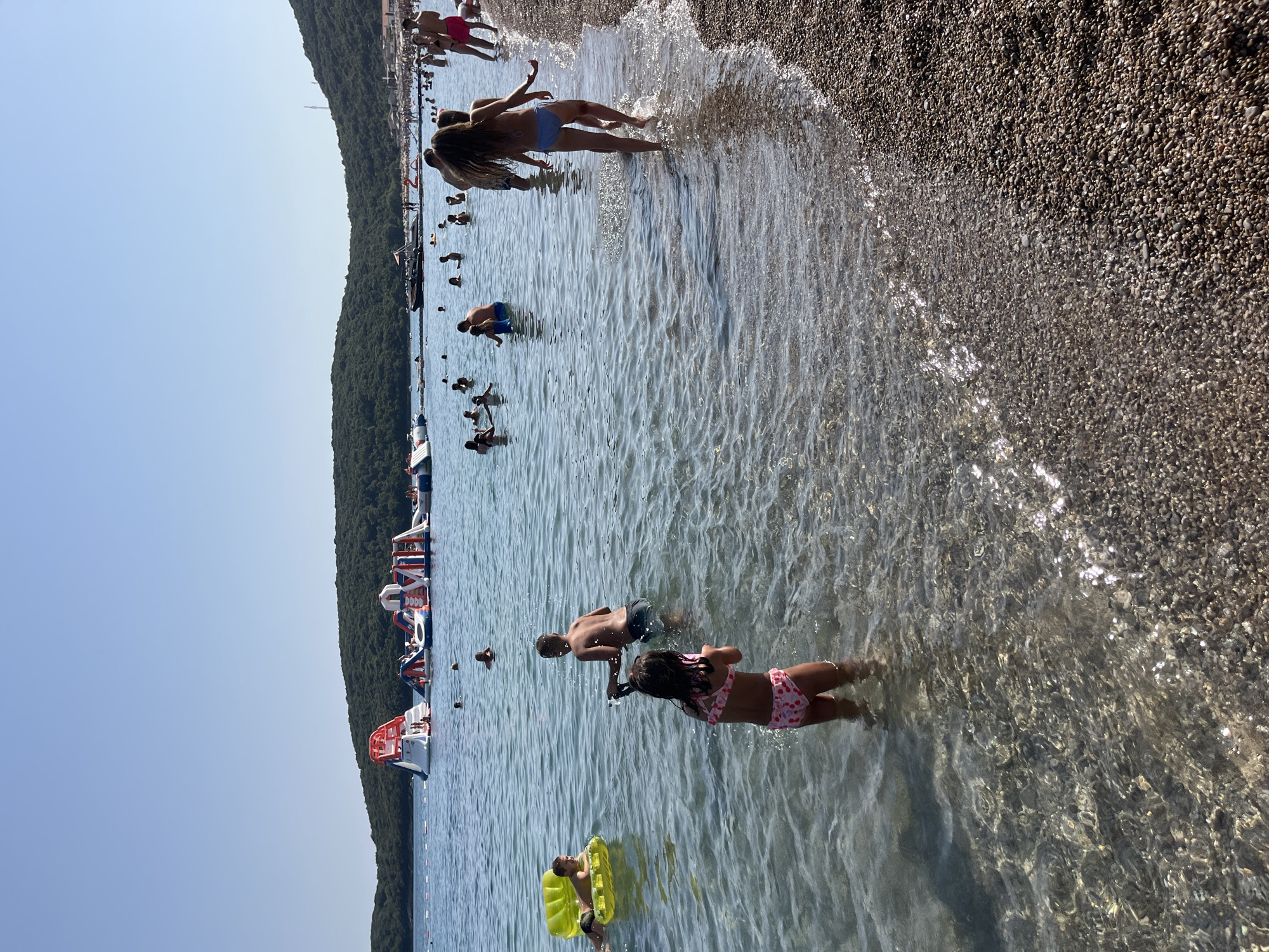 The image depicts a sunny day at the beach with numerous people enjoying the water and various inflatable water attractions. The water is clear and shallow near the shore, revealing a pebbled seabed. In the background, there's a lush green, tree-covered hillside that slopes down towards the sea, suggesting a coastal or bay location. On the water, several large, colorful inflatable structures, including slides and climbing walls, are visible, indicating a water park or amusement area. People of all ages are engaged in activities such as swimming, wading, and playing on the inflatables. Some individuals are closer to the shore, while others are further out in the water. The overall scene conveys a lively and recreational atmosphere typical of a summer beach outing. The location is identified as Prijevor, Montenegro.