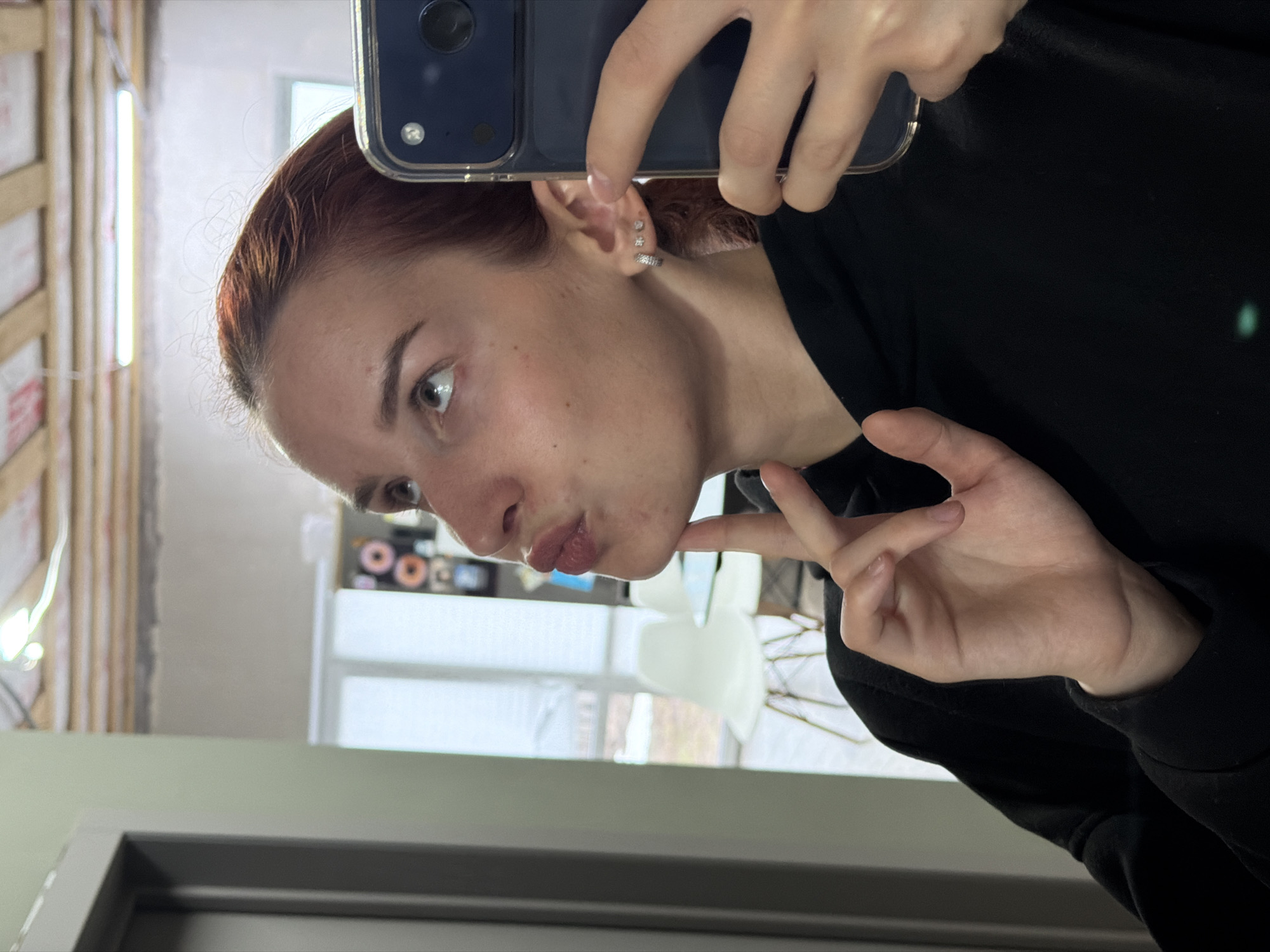 A woman with auburn hair, wearing a black hooded sweatshirt, is taking a selfie with a blue smartphone. She is pouting her lips and has her index finger touching her chin. She has multiple piercings in her ear. The background shows a room with a window and some interior furnishings including white chair with metal legs and shelves, and building materials leaning against a wall. The scene appears to be indoors. The general location is not possible to establish by the image.
