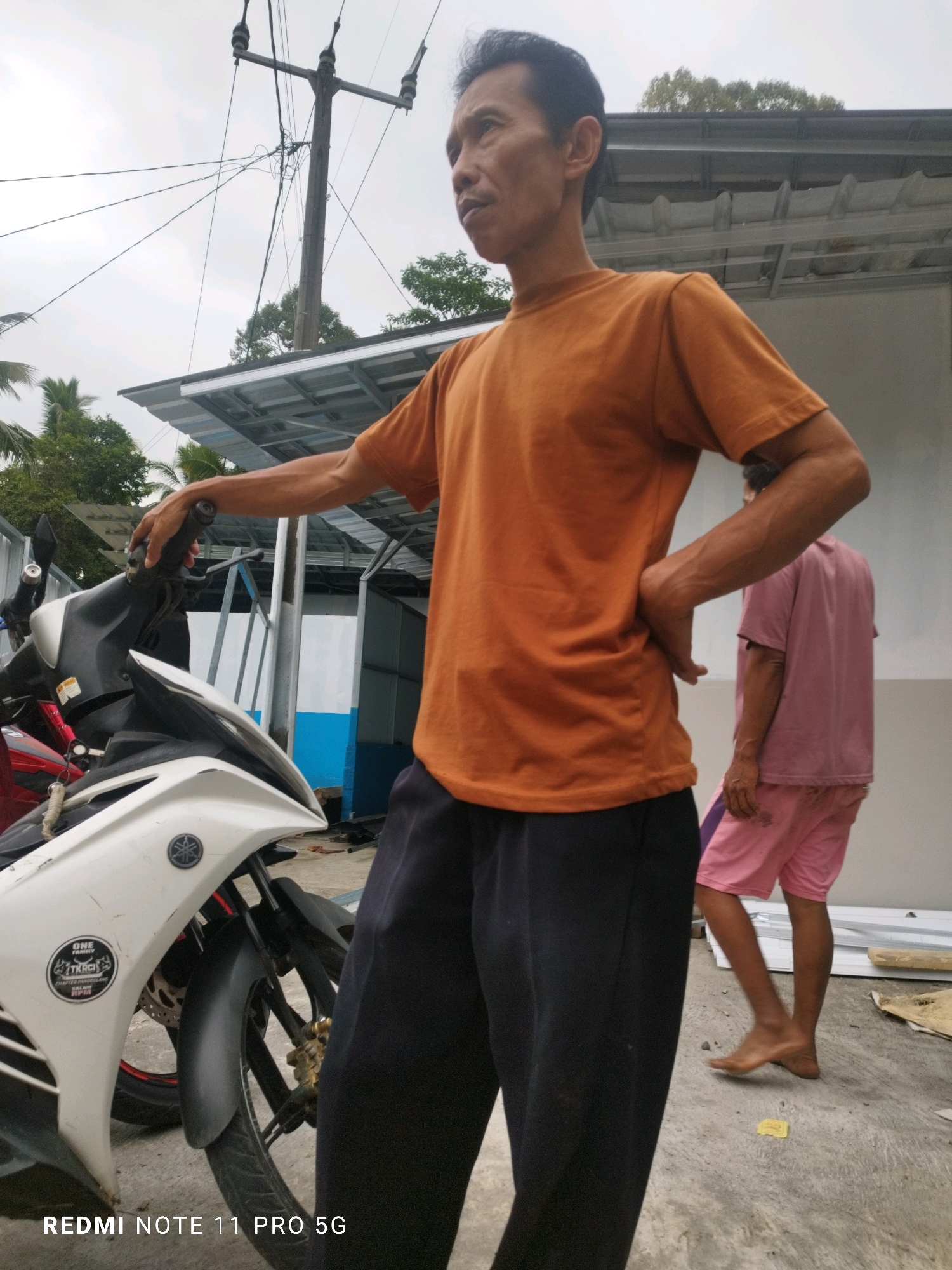 In Sindangkarya, Indonesia, a man with dark hair and a light complexion stands outdoors next to a white motorcycle. He is wearing an orange t-shirt and dark pants, with his right hand resting on the motorcycle's handlebar and his left hand on his hip. Behind him, another person is visible, wearing a pink t-shirt and pink shorts, walking away from the camera. The environment appears to be an outdoor construction or renovation area, with a grey concrete floor, a metal roof structure, and electrical wires and poles in the background. The sky is overcast.