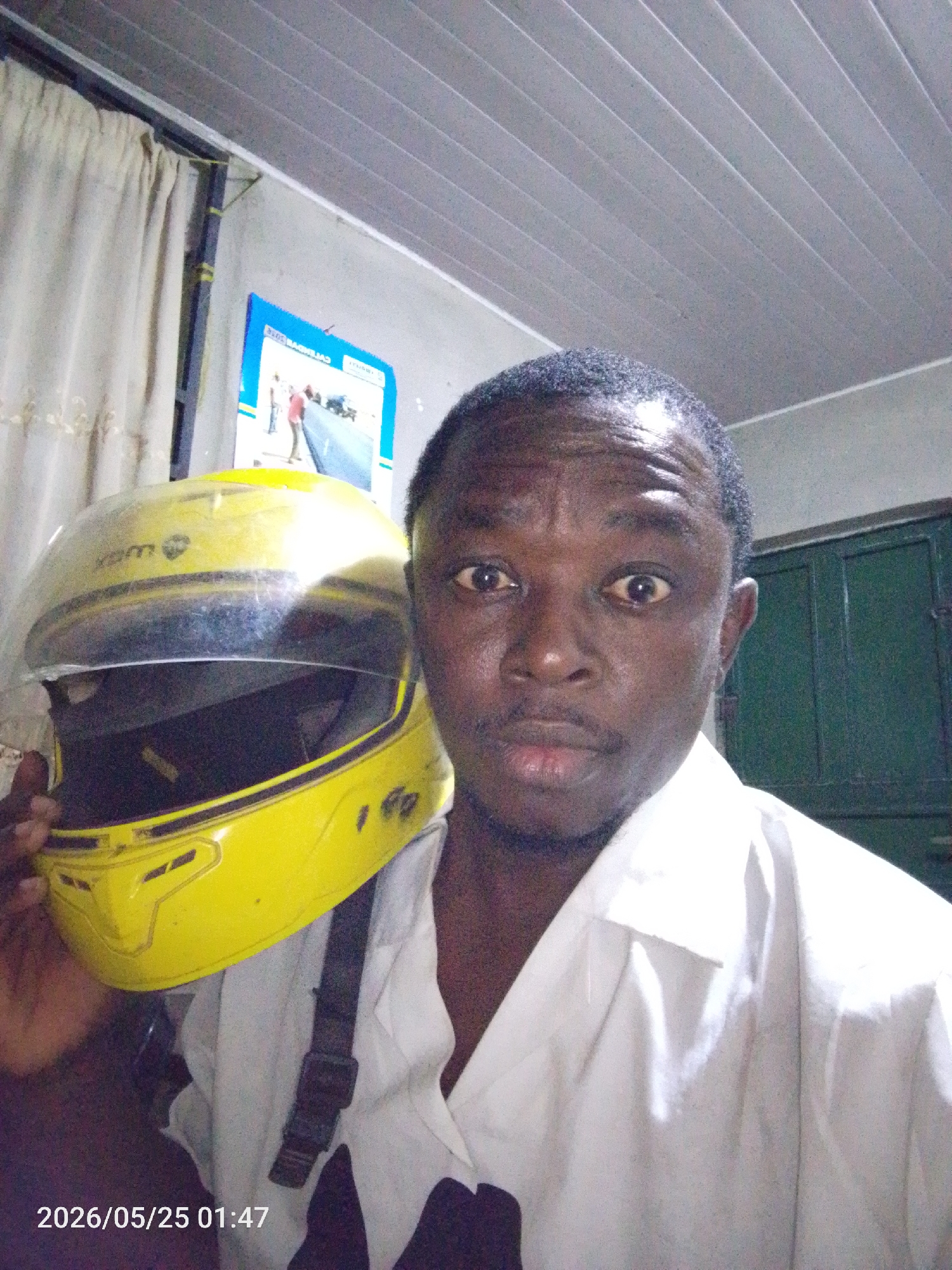 A man is holding a yellow helmet. He is wearing a white shirt. Behind him are curtains, a poster on the wall, and a door. The bottom of the image has the date "2026/05/25 01:47." Without additional information, it's impossible to confirm the location in Lokoja, Nigeria.