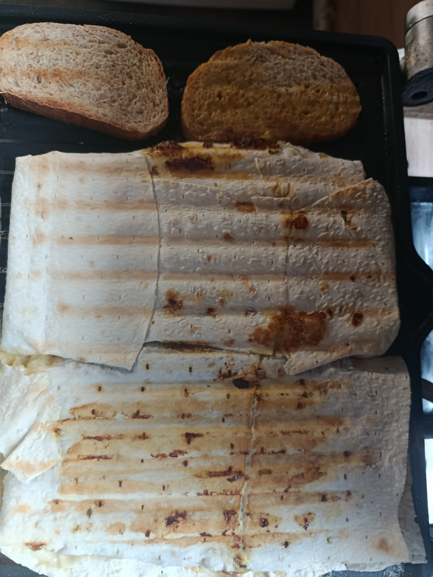 The image displays a close-up view of two grilled wraps and two slices of bread on a dark, textured surface, possibly a grill or stovetop. The wraps, made of thin, white flatbread, show distinct grill marks and some browning. The bread slices, one lighter and one darker, are also positioned on the surface. The scene is illuminated from above, casting subtle shadows. There are no people or other objects visible that would indicate a specific urban location.
