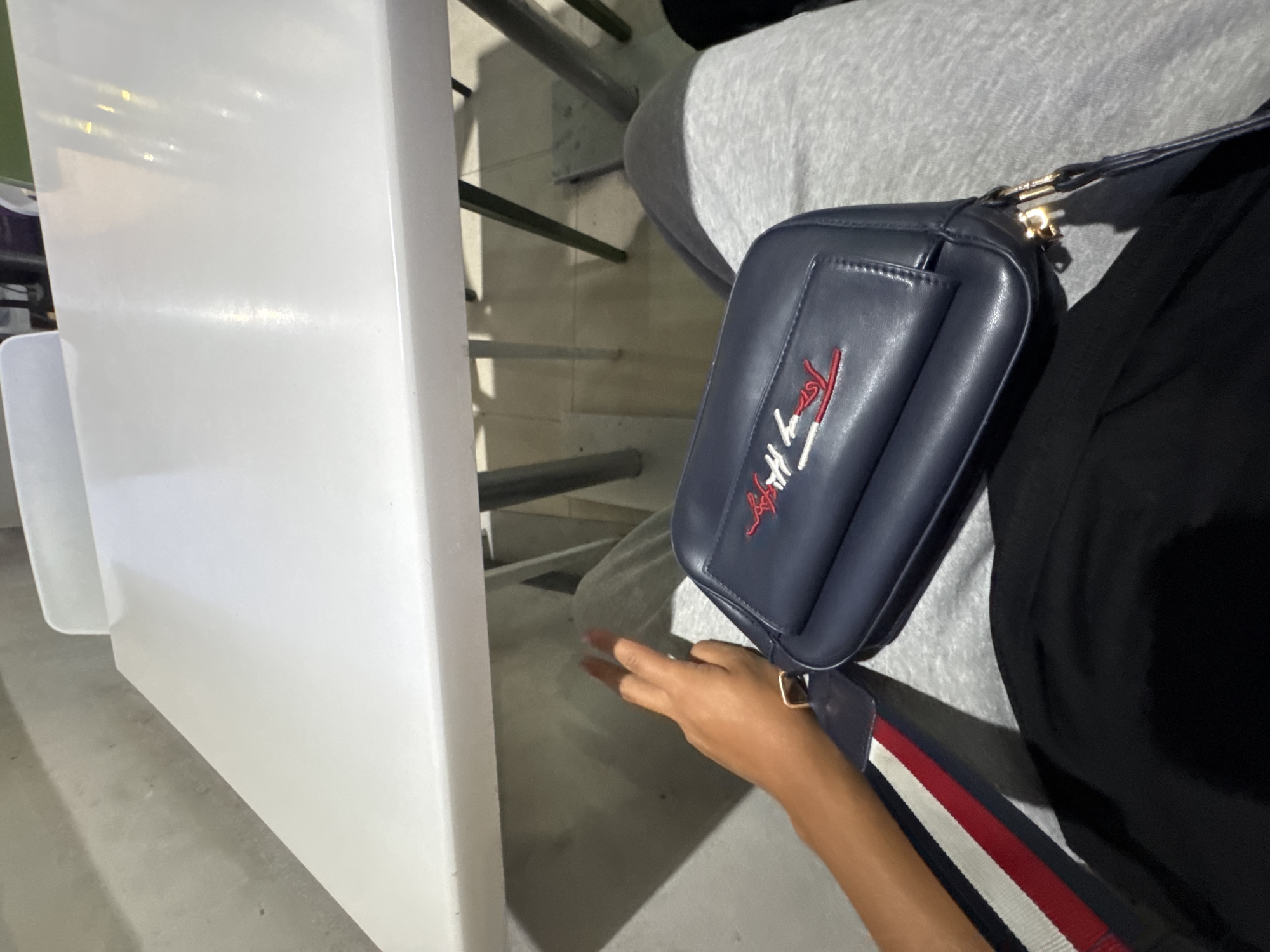 The image provides a close-up, slightly overhead view of a person seated in a public or semi-public setting in Yerevan, Armenia. The individual is partially visible, wearing grey trousers and a black top. A navy blue cross-body bag, featuring the "Tommy Hilfiger" logo embroidered in red and white, rests on their lap or beside them. The bag's strap, visible across their arm, is navy blue with prominent red, white, and navy stripes. The person's left hand and forearm, displaying light brown skin, are resting near the bag.
To the left of the frame, a tall, white barrier or partition dominates the foreground, with a smaller white object visible at its base. In the background, a concrete floor and a series of horizontal metal railings are visible, suggesting a structured environment such as a stadium, public transport station, or an indoor venue with tiered seating. The lighting is dim, indicating an indoor setting or evening/nighttime conditions. No specific activity is depicted beyond the person being seated. The only visible text is the "Tommy Hilfiger" brand name on the bag.