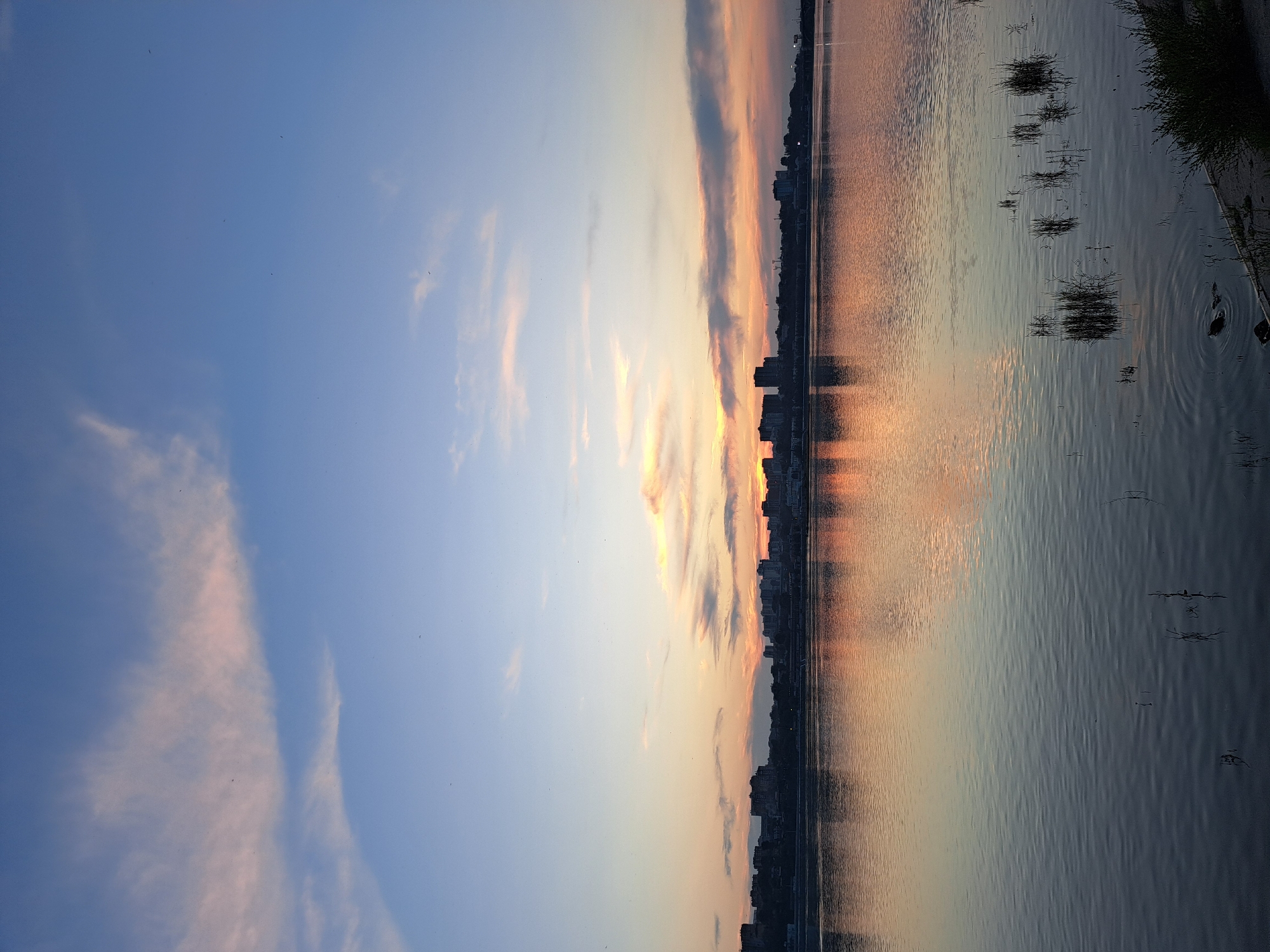 This is a landscape photograph of the Angara River in Irkutsk, Russia, at sunset. The sky is a gradient of blue and pink, with wispy clouds illuminated by the setting sun. A distant cityscape, characterized by silhouetted buildings, lines the horizon. The river's surface is calm, reflecting the colors of the sky and the city. Patches of dry grass and reeds are visible along the riverbank, and a few small birds are in flight. The overall atmosphere is serene and tranquil.