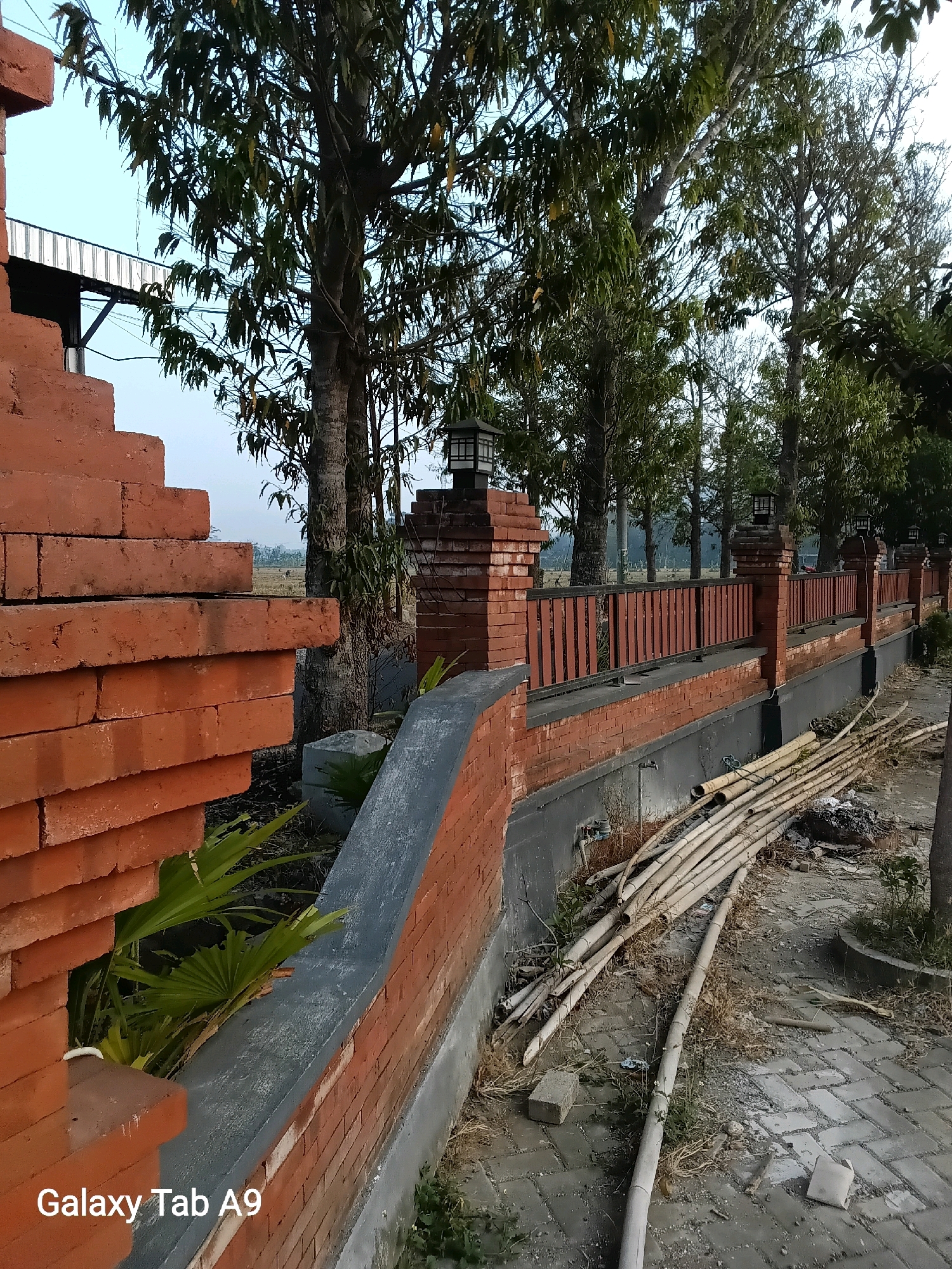 An outdoor view shows a brick wall and fence in Bendorejo, Indonesia. In the foreground, a stacked brick structure rises from the left of the frame. A brick wall with a gray cap extends horizontally across the middle ground, topped by a wooden slatted fence. Several trees with sparse leaves are visible behind the fence, and the sky is a pale blue. In the right foreground, a pile of bamboo poles lies on the ground next to a paved walkway. The scene appears to be a construction or renovation area.