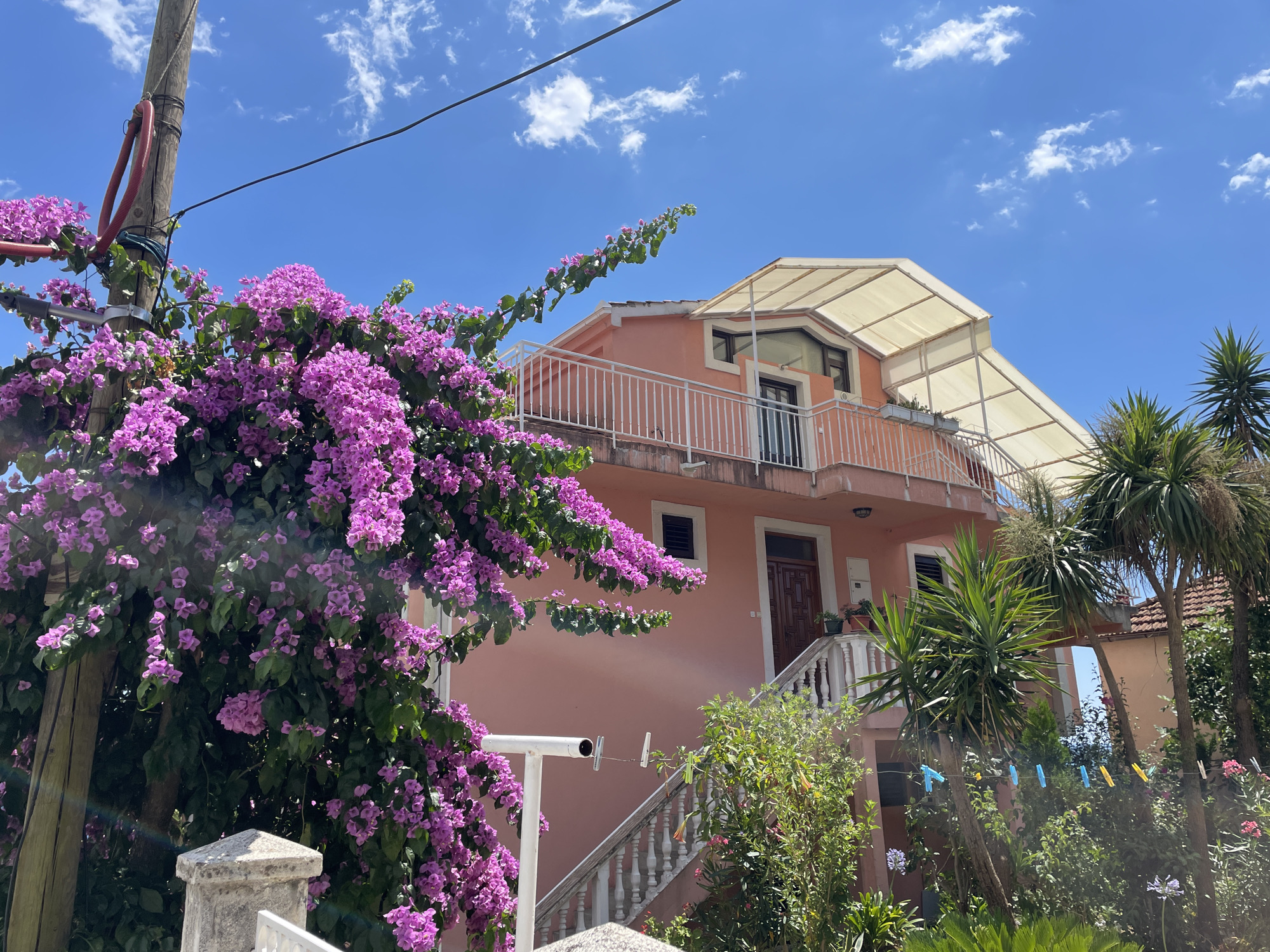 In Herceg Novi, Montenegro, a pink house is visible on a sunny day under a blue sky with scattered clouds. A vibrant bush of purple bougainvillea blooms in the foreground, partially obscuring a wooden utility pole and a stone pillar. The house features a white balcony with a railing and a large, cream-colored awning extending from the roof. An outdoor staircase with a white railing leads to the front door, which is made of dark wood. Various green plants and palm trees surround the property. A white security camera is mounted on a pole, and clothespins hang on a wire strung between two poles in the yard. The scene depicts a residential area with lush vegetation and a clear, bright sky, characteristic of a Mediterranean climate.