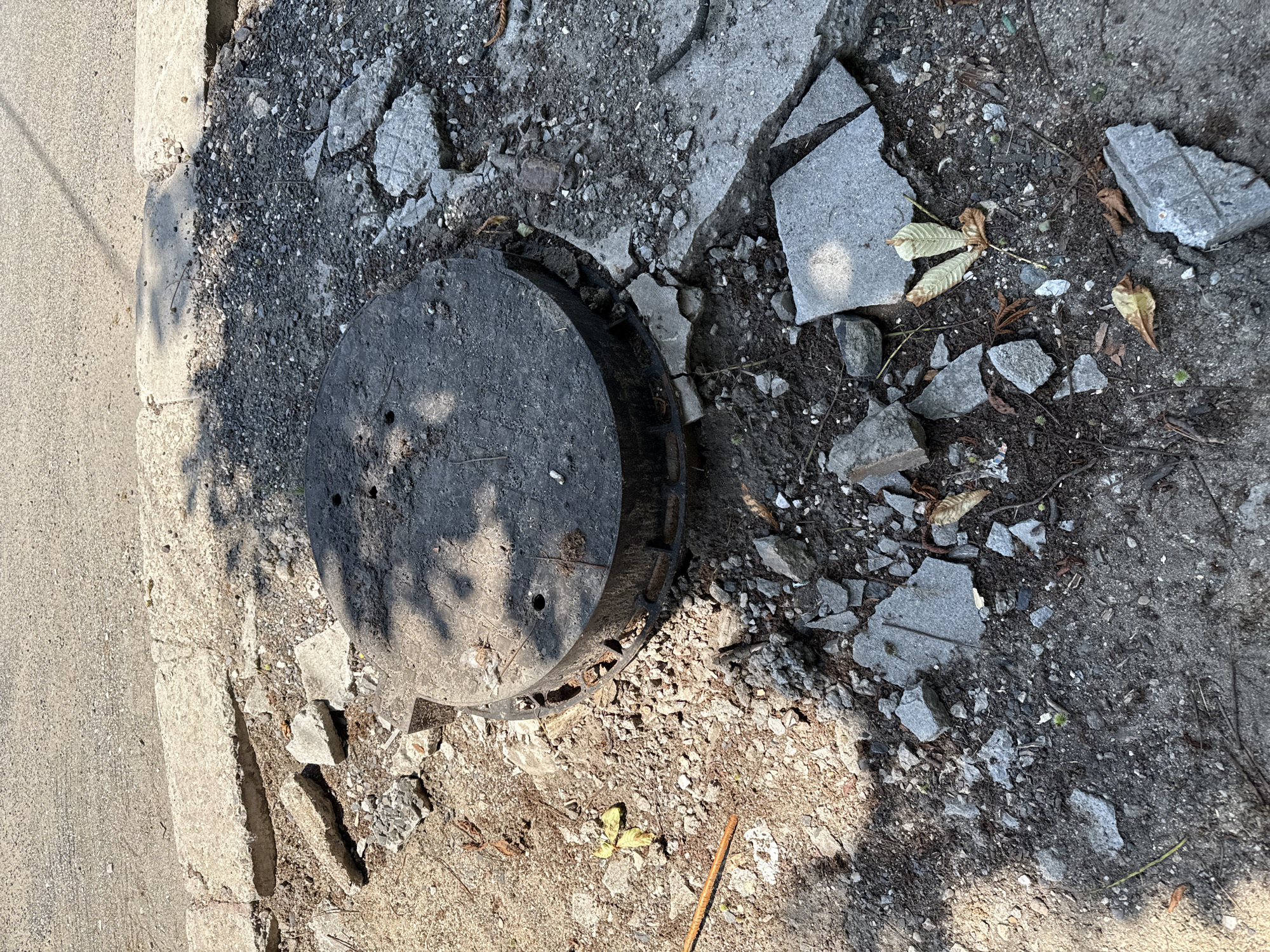 This forensic image depicts a severely degraded section of ground adjacent to a road in Kardzhali, Bulgaria, under direct sunlight. The central subject is a dark grey, circular manhole cover, likely constructed from concrete or a composite material, featuring two small circular perforations on its surface. The manhole cover is situated within a substantial area of severely fractured and disintegrated concrete, characterized by numerous fragments of varying sizes interspersed with dirt, gravel, and small stones.
Immediately surrounding the manhole's base, the concrete infrastructure exhibits significant breakdown and unevenness. Towards the upper portion of the image, a light grey asphalt road surface is visible, separated from the debris field by a broken and uneven concrete curb.
Scattered across the scene are organic debris, including several dried leaves, some resembling chestnut leaves, and small twigs. A rusted metal rod or rebar segment lies embedded in the debris in the lower-left area. Sparse, small green vegetation protrudes from the fractured ground. No individuals or active subjects are present, and no specific actions or interactions are depicted beyond the observable state of disrepair. Shadows, consistent with dappled sunlight passing through foliage, are cast across the manhole cover and the surrounding ground.